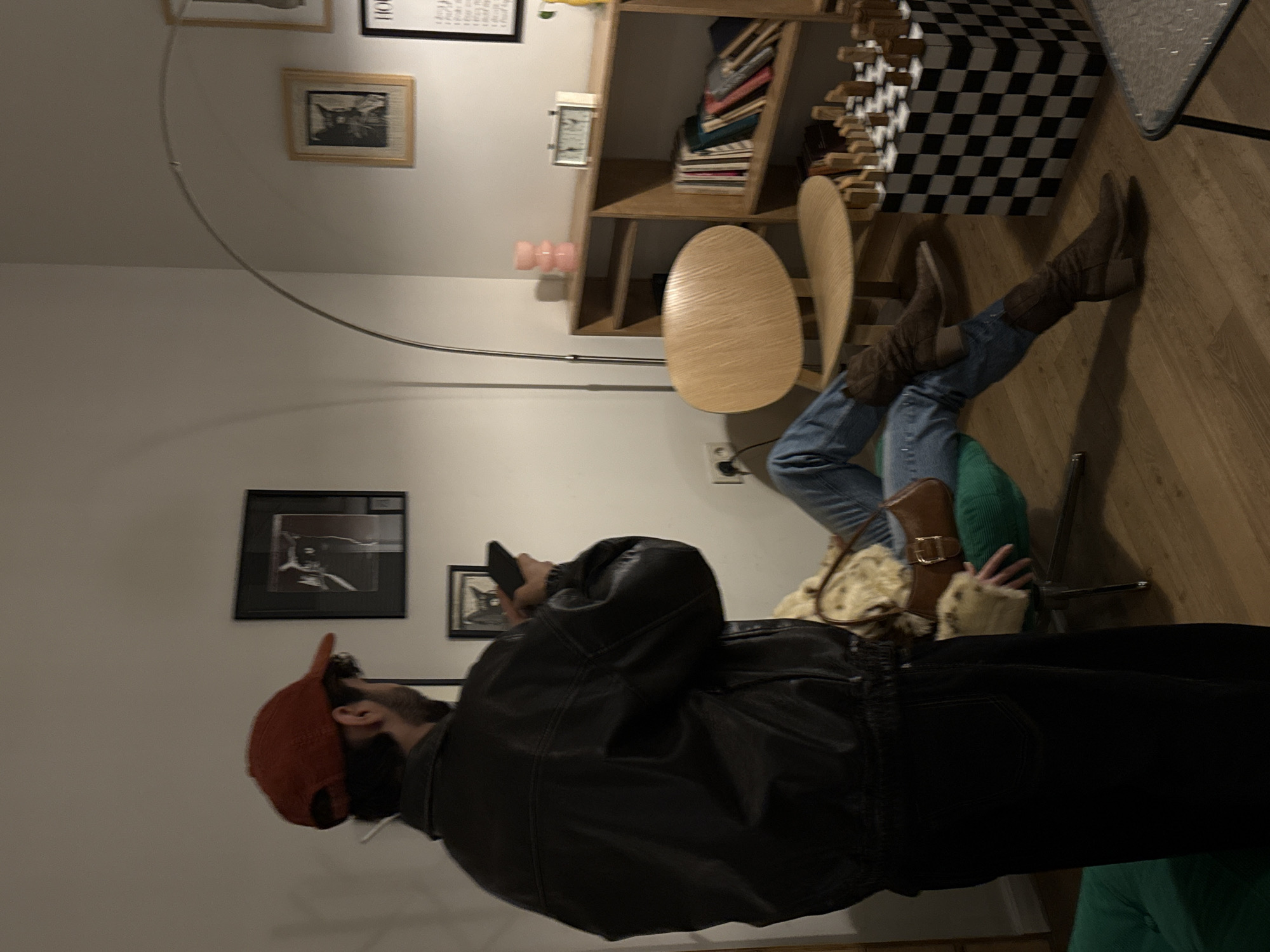 A man wearing a black leather jacket and a red cap is standing in a room, holding a mobile phone. The room has hardwood floors and is furnished with a light wood table and chairs. A person wearing blue jeans and brown boots is seated. There are framed pictures on the walls. The exact location in Yerevan, Armenia, cannot be determined from the image.