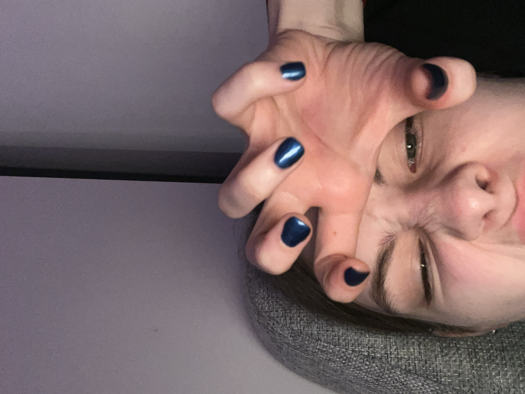 A person's face and hand are in focus, angled as if the person is looking down at the viewer from above. The person has fair skin, with blue nail polish on their fingers. They are squinting with a slight frown, and are resting on a gray textured surface. The background consists of a gray and white wall, possibly a ceiling. The photograph provides no information to confirm or deny the location being in the city of Saint Petersburg, Russia.