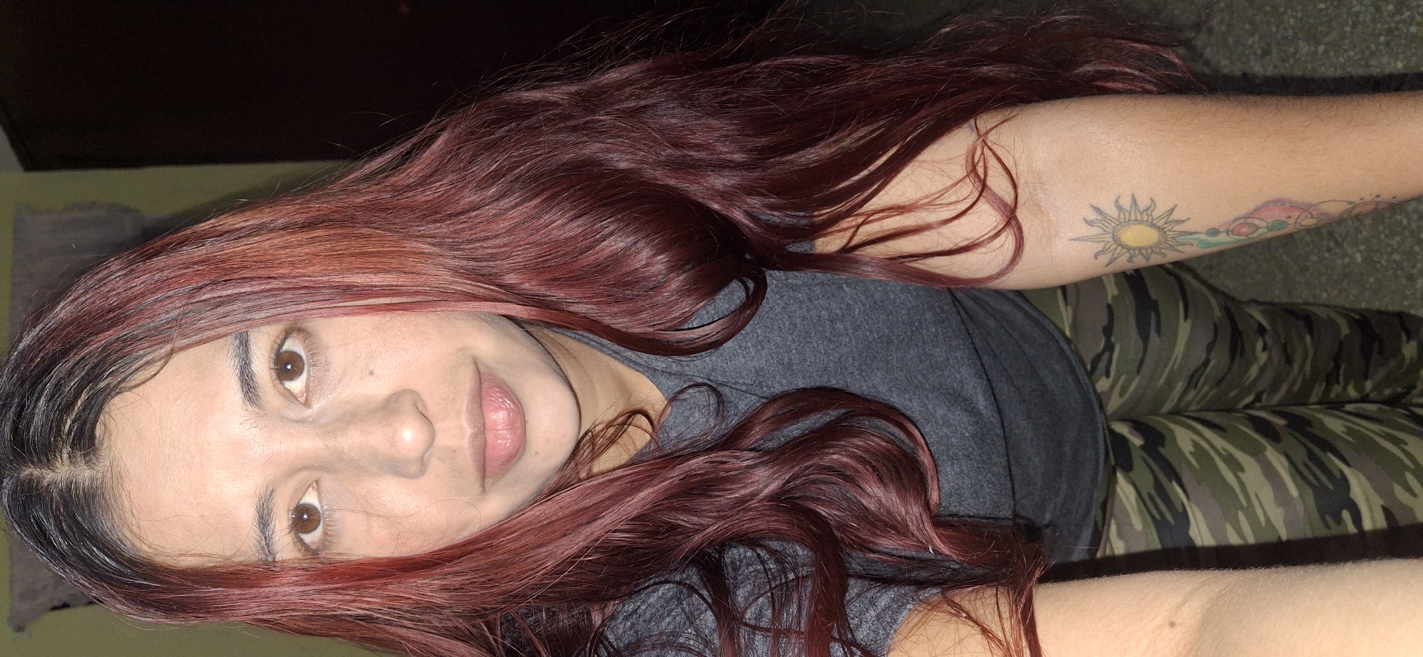 A woman with long, dark red hair is lying down. Her eyes are brown, and her skin tone is light. She is wearing a gray t-shirt and camouflage pants. There is a tattoo of a sun and other colorful designs on her left arm. The background is dark and indistinct. There is no location information for the city of Tinaquillo, Venezuela in the image.