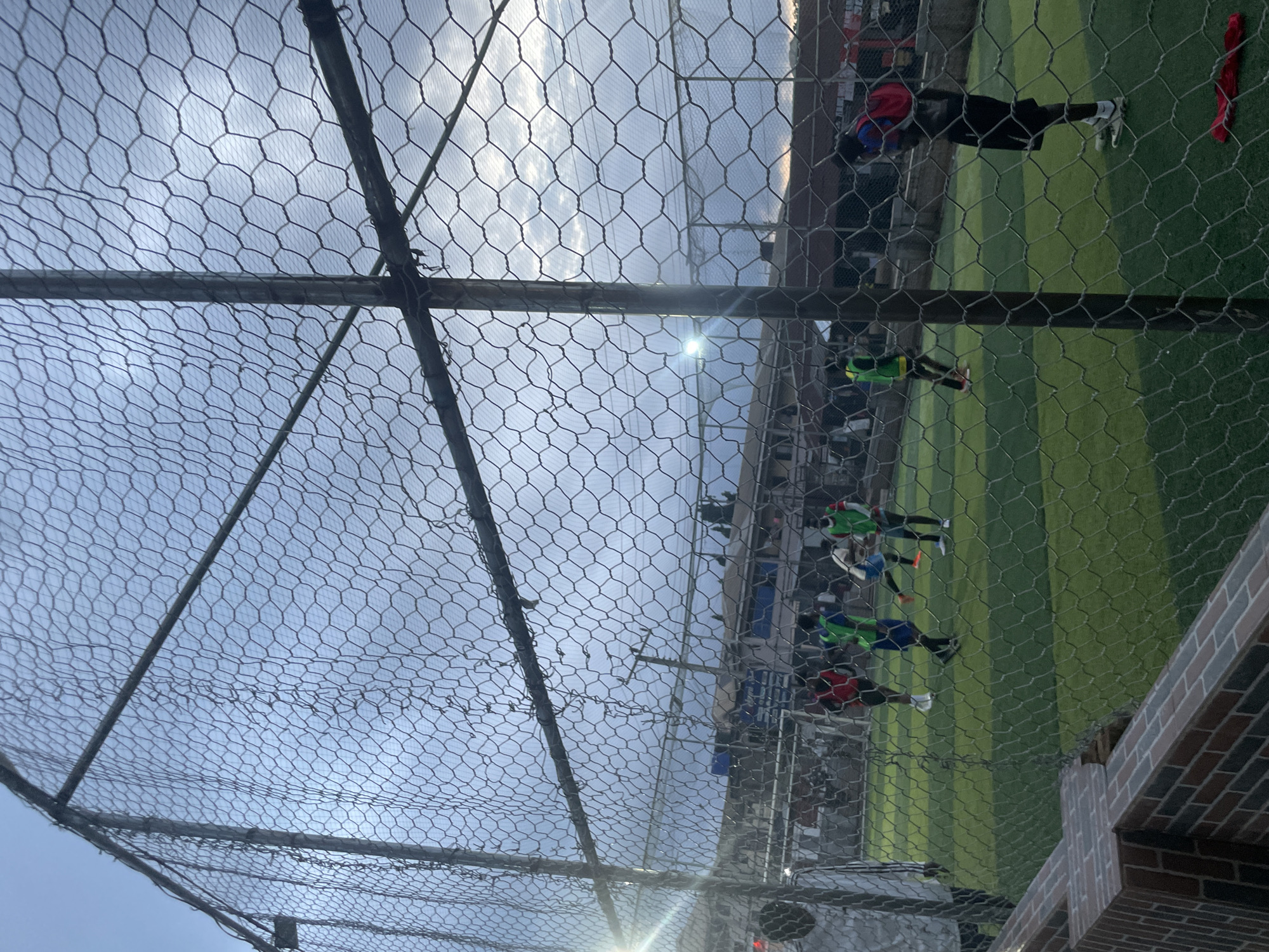 This image depicts an outdoor artificial turf soccer field in Ihiagwa, Nigeria, viewed through a wire mesh fence. The sky is overcast with a hint of sunlight breaking through. Several individuals are present on the field, engaged in a soccer game. Two individuals in red and green vests appear to be actively playing soccer. Another person in a red vest is standing on the right side of the field, facing away from the camera. The surrounding area shows low-rise buildings and some vegetation. The foreground includes a brick wall and the prominent wire mesh fence that obstructs the view of the scene. A bright light, possibly a floodlight, is visible on the left side of the field.
