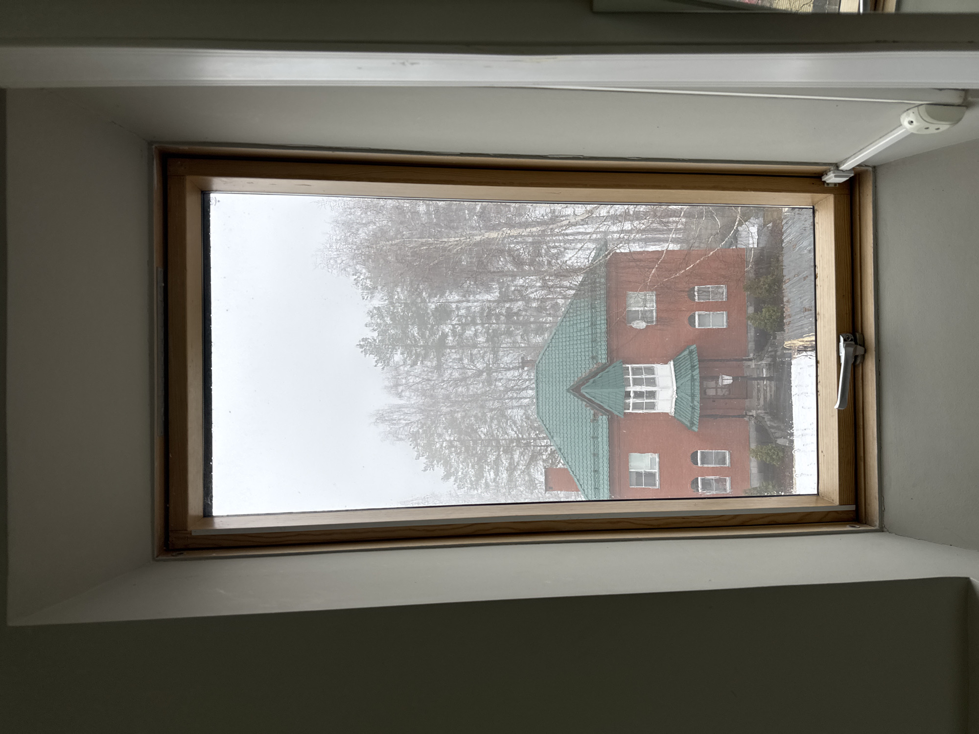 A window, framed in light-colored wood, looks out on a brick building with a green roof. The building has white-framed windows. Trees are visible between the window and the building. It appears to be a snowy or foggy day. The window itself is mounted on an angled wall and has a white window handle on the right. The location is unknown.