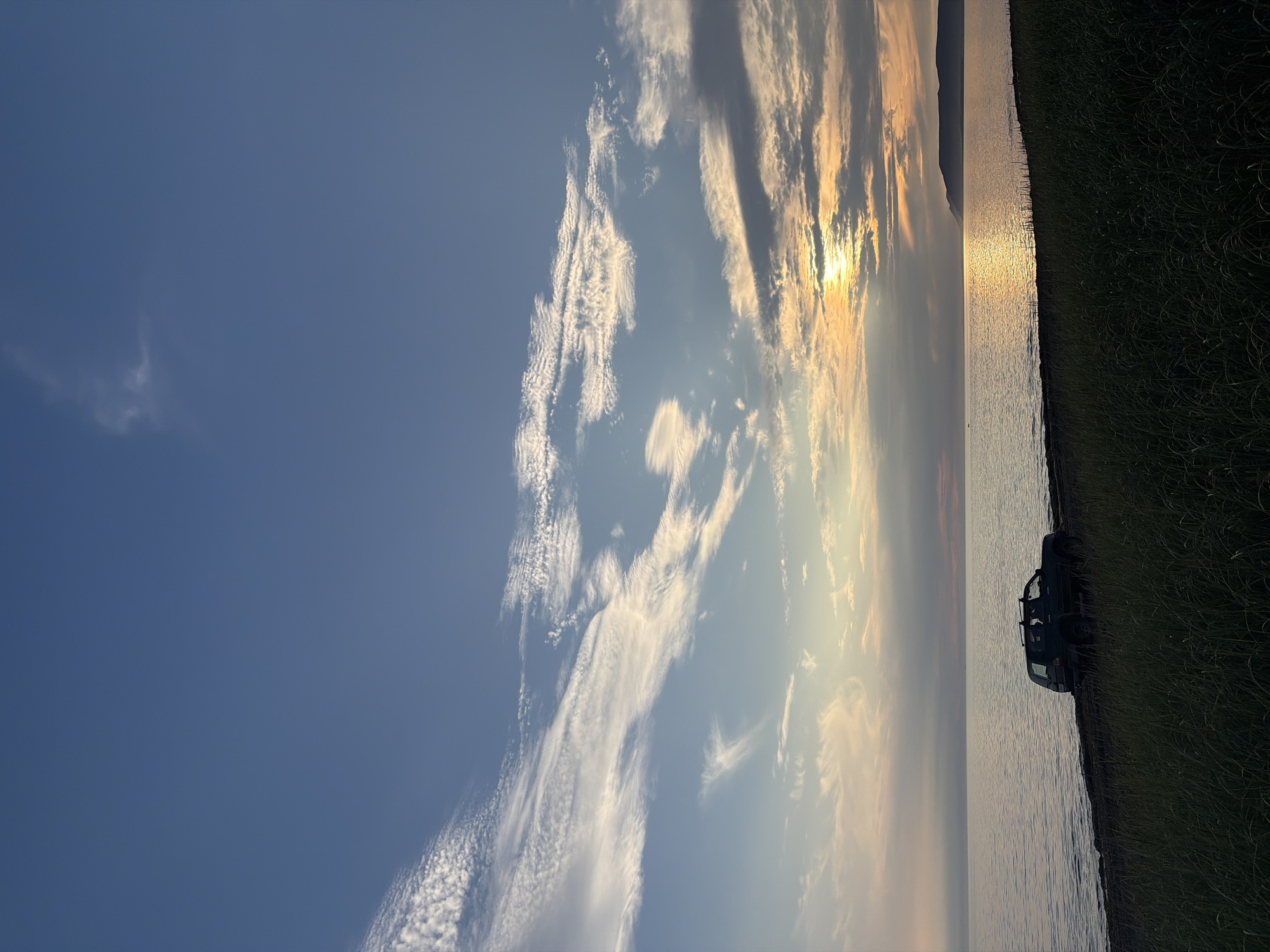 A dark SUV is parked on the shore of a body of water. The water is calm and reflects the sky, which is filled with wispy clouds and a setting sun. The foreground is dominated by tall grass. The scene appears to be in Ozerskoe, Russia.