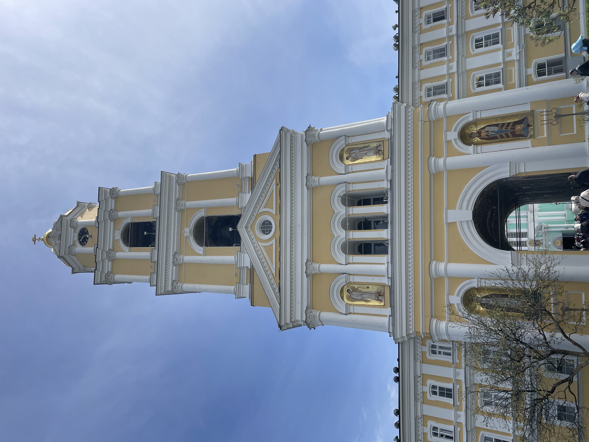 Captured from a low angle looking up, the image features a tall, yellow and white building with a multi-tiered tower set against a bright, clear sky. The building exhibits classical architectural details including columns, arches, and ornamentation. Ornate mosaics or religious figures are set into niches along the facade. The building appears to be a religious structure, possibly a church or cathedral. Bare branches of a tree are visible in the bottom right corner. Part of the building appears to have an archway that leads to a building with green windows. The location is Unknown, Unknown.