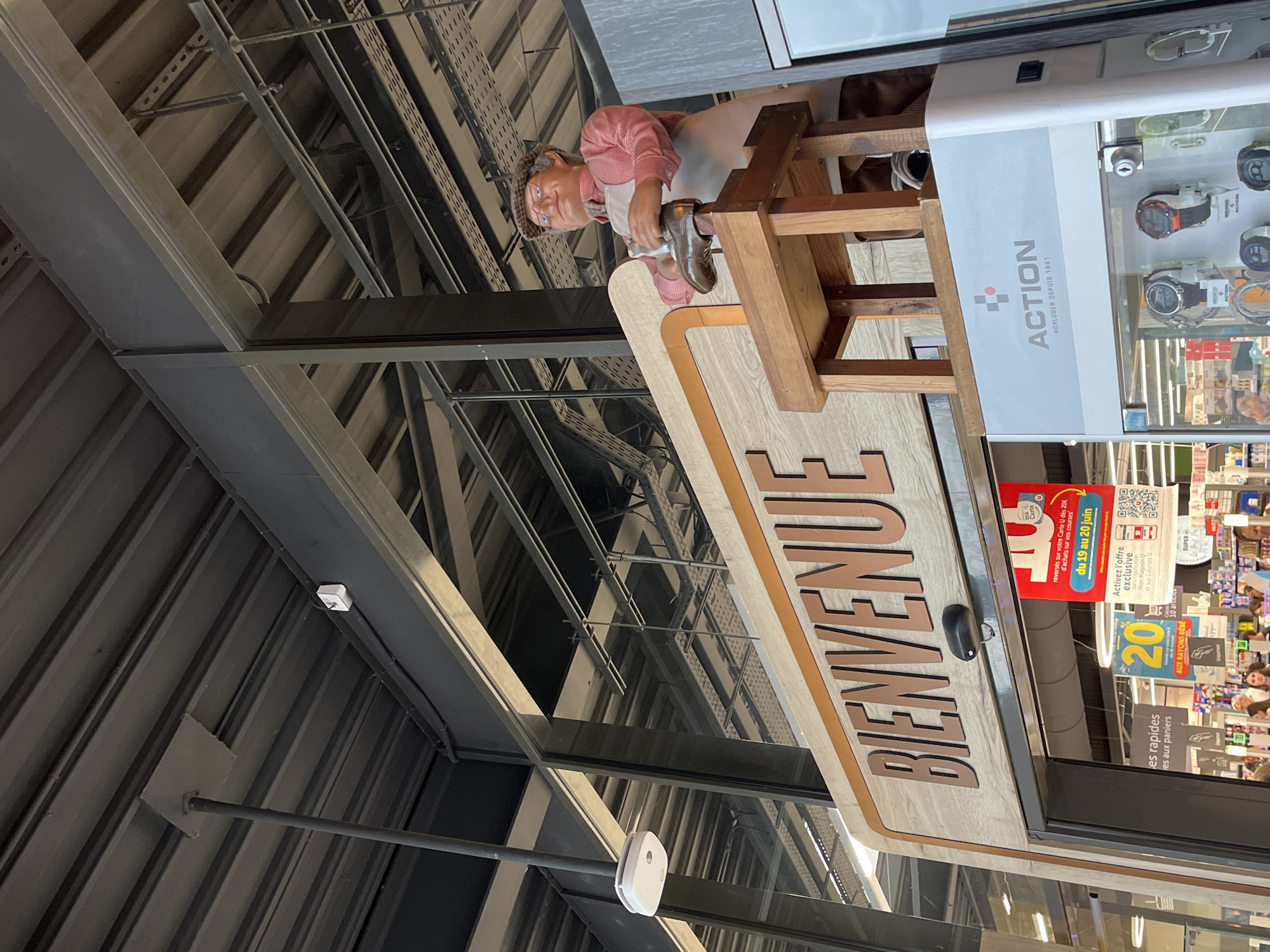 The image shows the entrance to a store, likely a supermarket, in Le Port-Marly, France. A large sign with the word "BIENVENUE" (Welcome) is prominently displayed above the entrance. To the right of the sign, a life-sized mannequin of a shoemaker is positioned on a wooden stand, holding a shoe. Below the sign, a display case with watches is visible, featuring the "ACTION HORLOGER DEPUIS 1961" branding. The interior of the store, with shelves and shoppers, can be seen through the entrance. The overall environment suggests a commercial setting, possibly a hypermarket or a large retail store. The ceiling is industrial, with exposed metal beams and ductwork.