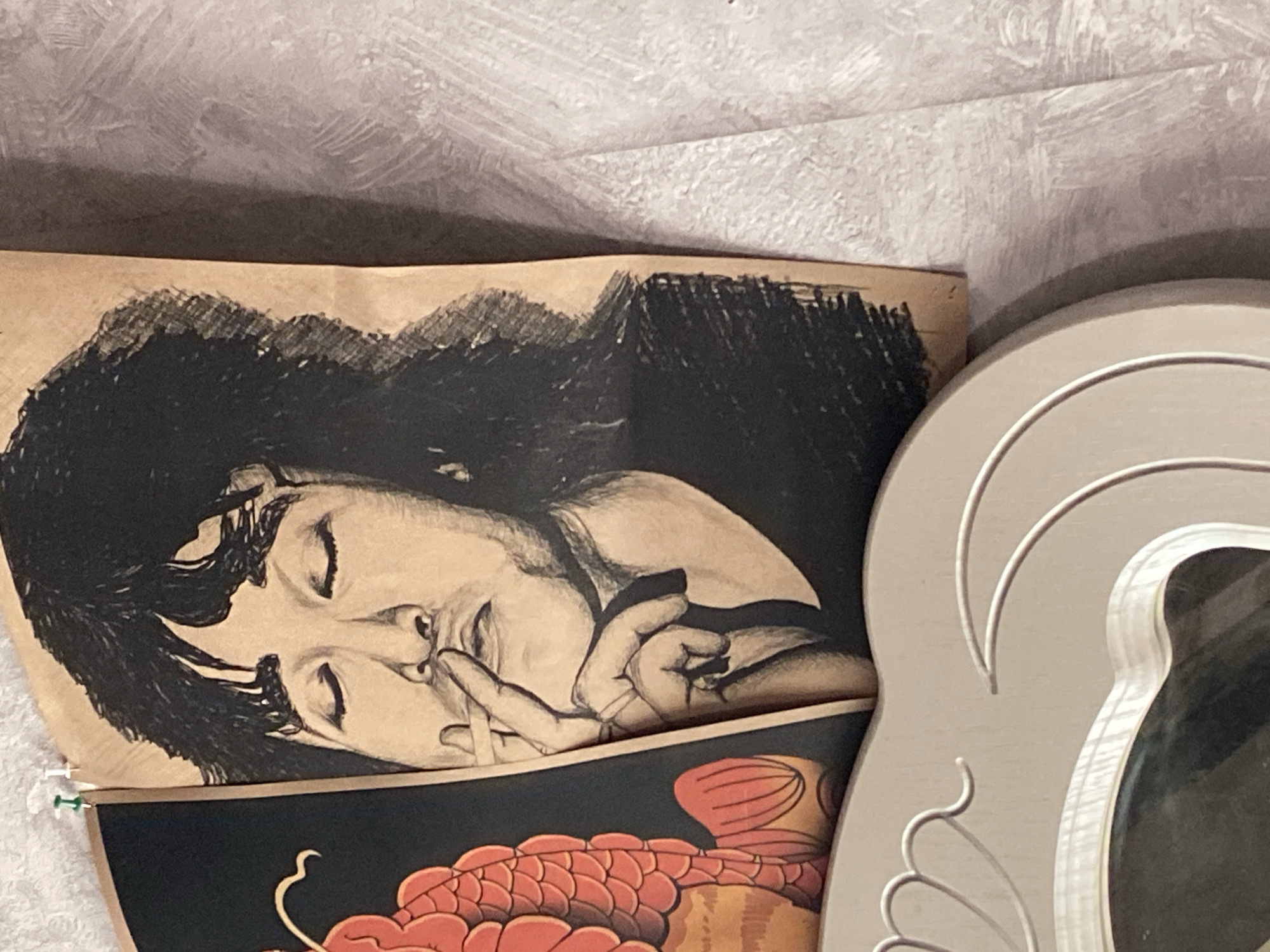 The image shows a drawing of a person with long, dark hair, eyes closed, holding a cigarette to their lips. The drawing is on a brown paper, attached to a wall with a pushpin. To the left of the drawing is a portion of a black and red illustration of what appears to be a fish. In the foreground, a portion of a light-colored, ornate frame is visible. The drawing and illustrations are hung on a textured wall. The specific location in Shakhtinsk, Kazakhstan is not provided.