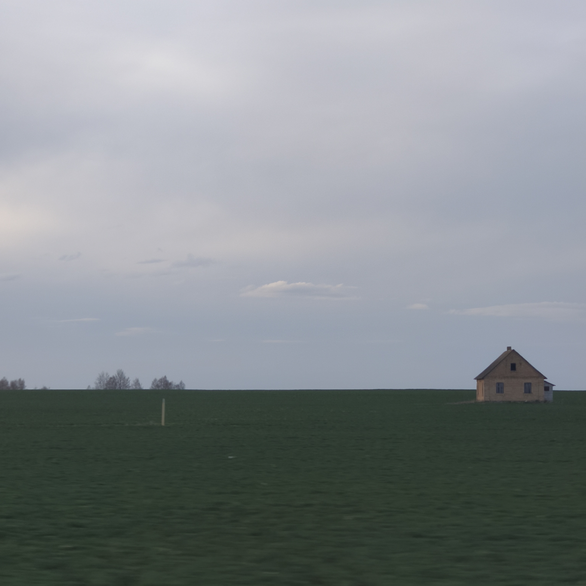 The image shows a solitary house in a field under a cloudy sky. The house is small, likely a single-story structure with a gabled roof and a chimney. The field is green, and the sky is overcast with shades of gray. A few bare trees are visible on the horizon in the distance on the left. There is also a single wooden post. The location is likely within the vicinity of Лунненскі сельскі Савет, Belarus, given the information provided.