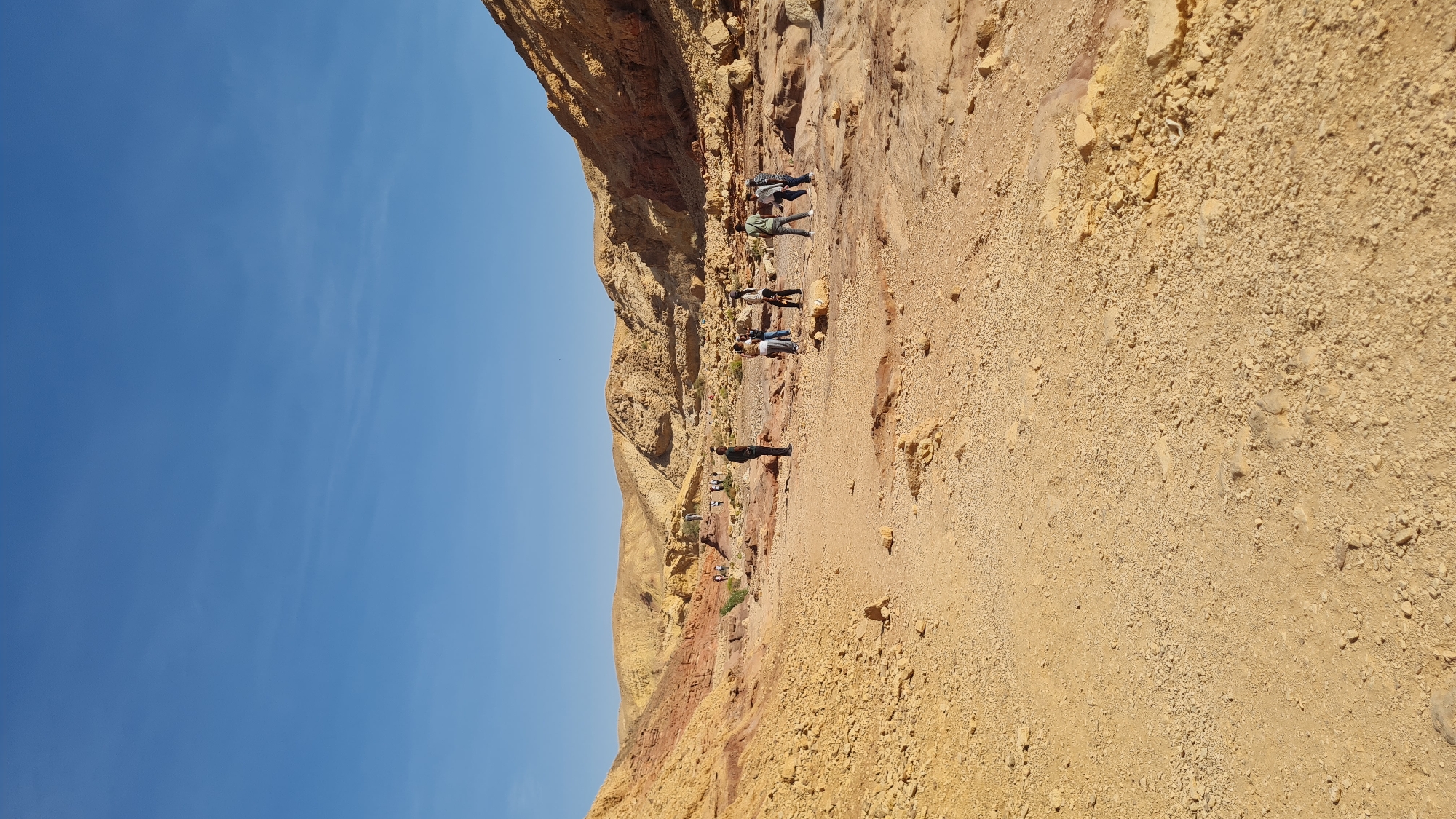 The image shows a rocky, desert-like landscape in the city of Hevel Eilot Regional Council, Israel. A group of people are standing near a rock formation with different layers of color, ranging from yellow to red. The sky is clear blue. The ground is sandy and rocky.