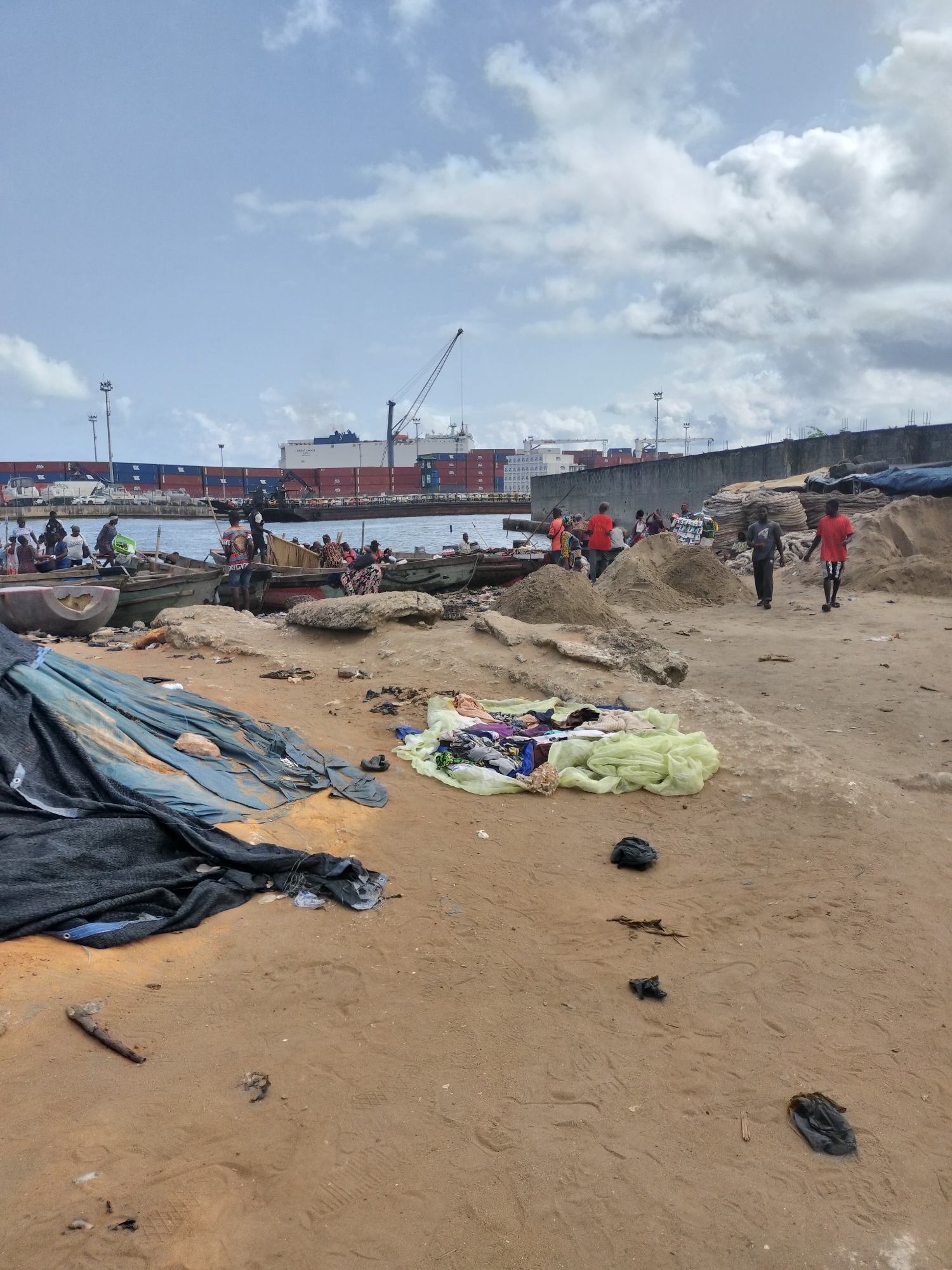 This full-shot image depicts a busy, informal waterfront scene in Alaiyabiagba, Nigeria, under a partly cloudy sky during daytime. The foreground is sandy ground littered with a large dark blue/black tarpaulin, a pile of lighter green fabric with various clothes on it, and scattered debris including a piece of wood and some dark objects resembling plastic bags or shoes. Footprints are visible in the sand.
In the midground, numerous people are gathered around several traditional wooden boats (pirogues) partially pulled ashore or moored in the water. Individuals are seen getting into or out of the boats, and some appear to be loading or unloading goods. Many people are dressed in casual attire, with some wearing bright red or orange shirts. Piles of sand or earth are scattered across the sandy area where people are walking. Two men are walking away from the boats on a sandy path towards the right of the frame.
The background reveals a working port. Large container ships are docked, with visible shipping containers, some bearing the "MSC" logo. One white cargo ship clearly displays the name "GREAT EAGLE" on its side. Industrial cranes and port infrastructure, including a concrete barrier and tall light poles, are visible along the waterfront. The overall atmosphere suggests a bustling hub of trade and maritime activity.