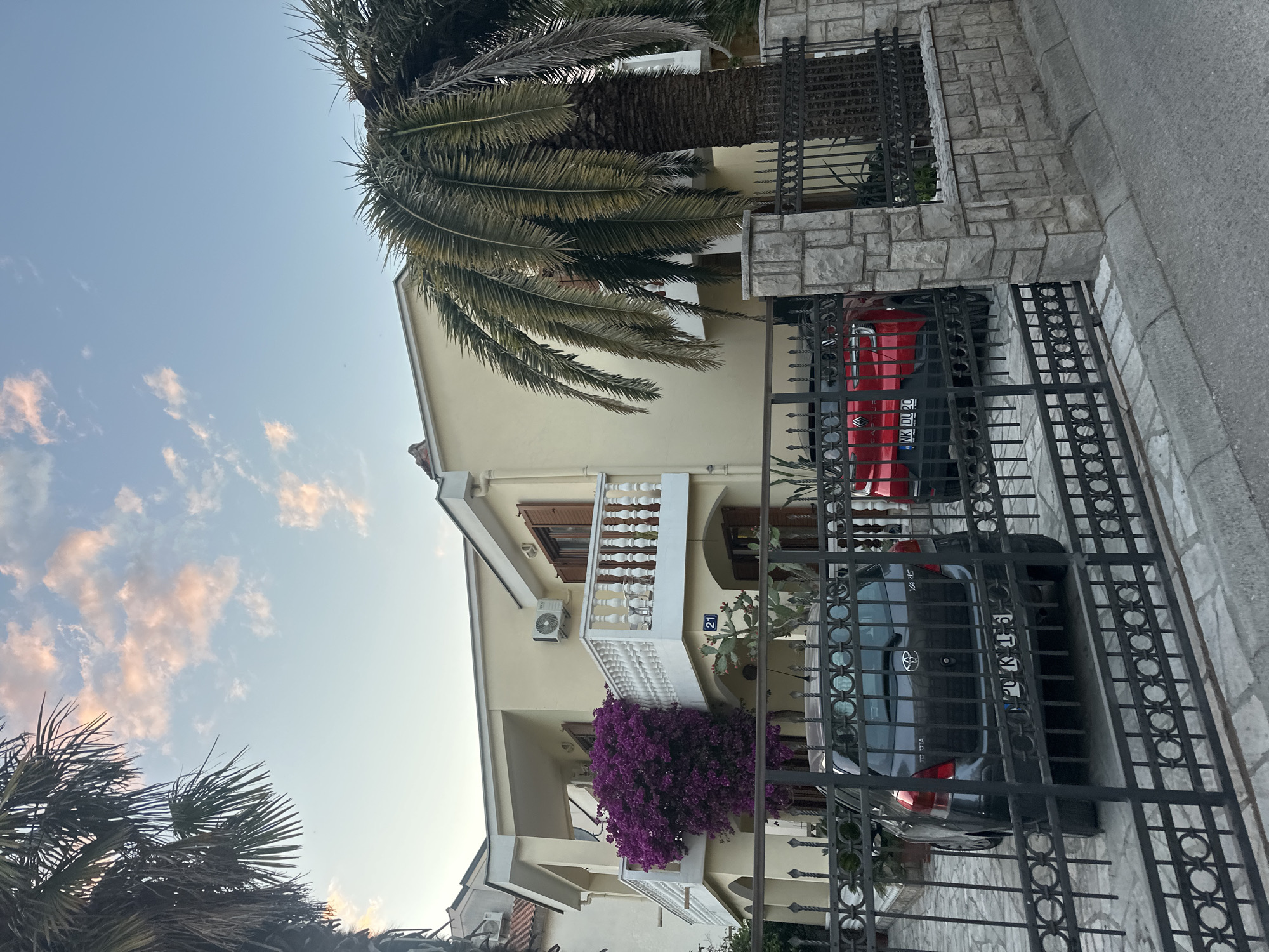 A medium shot of a two-story building in Budva, Montenegro, taken at ground level from outside a black metal fence. The building is painted a light yellow color and has two cars parked in front, one red and one gray. A large palm tree is visible behind the building and the fence, and a purple flowering vine grows over the lower balcony of the building. A balcony on the second story has a small sign with the number 21. The sky is light blue with pink-tinged clouds.