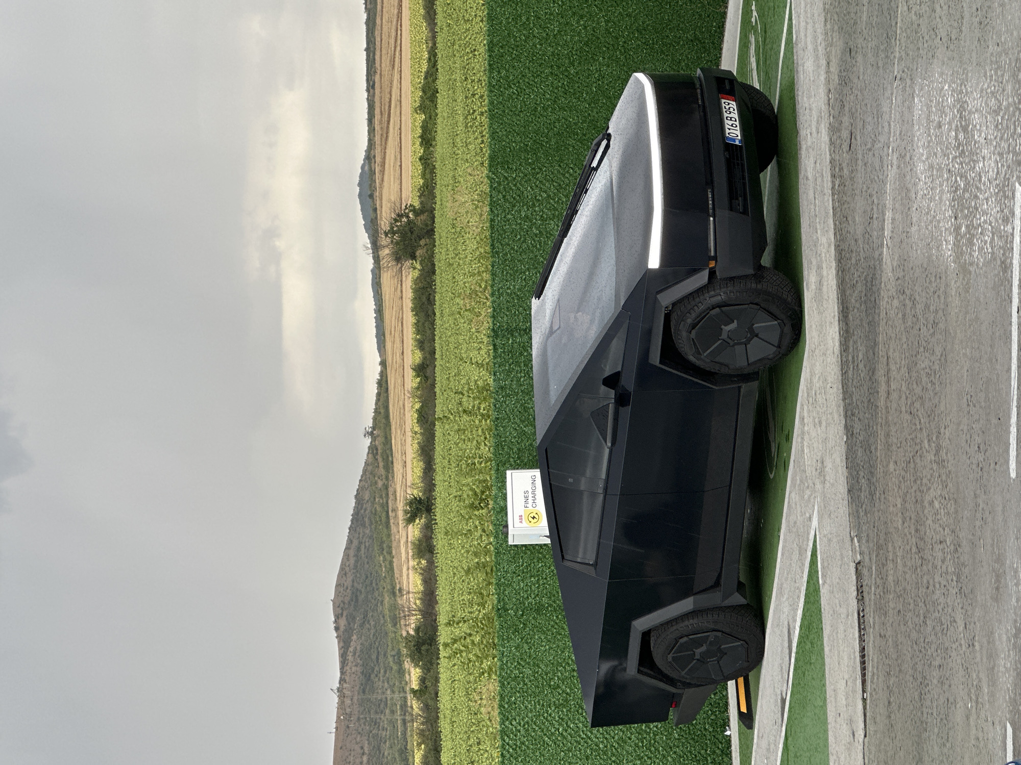 In Polsko Padarevo, Bulgaria, a black Tesla Cybertruck is parked in a charging bay, indicated by the green turf and white lines. Behind the vehicle, a green, textured fence separates it from a field of green crops, which extends to the horizon. Beyond the field, rolling hills with sparse trees and a cloudy, overcast sky form the background. A white charging station sign with the ABB logo and "FINES CHARGING" text is visible on the fence. The truck's distinctive angular design, LED light bar, and license plate "016 B 959" are clearly visible. The ground surface is a mix of asphalt and the green turf of the charging bay, with some evidence of wetness.