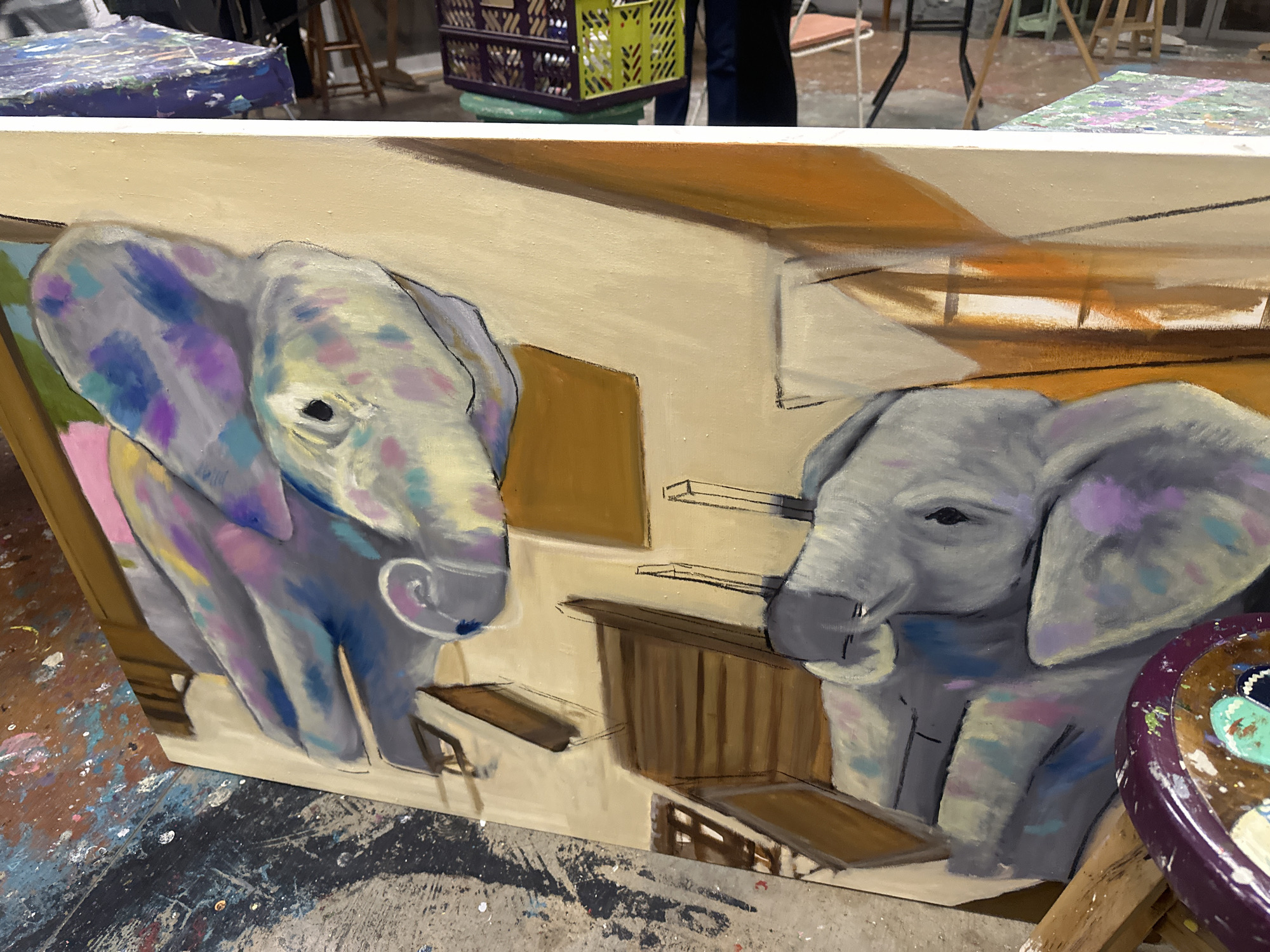 A painting of two elephants is in the foreground. The left elephant is larger and painted with more color variation, including blues, pinks, purples, and yellows. The right elephant is painted primarily in grays and purples. Both elephants are positioned within an abstract, geometric interior, painted in shades of beige, brown, and orange. The painting is on a canvas and appears to be resting on a surface covered in paint splatters, indicative of an artist's studio. In the background, there are other objects, including a crate and a table covered with painted materials. The environment is cluttered and suggests a creative workspace. The location, San Bernardino Tlaxcalancingo, Mexico, is not evident from the visual content of the image.