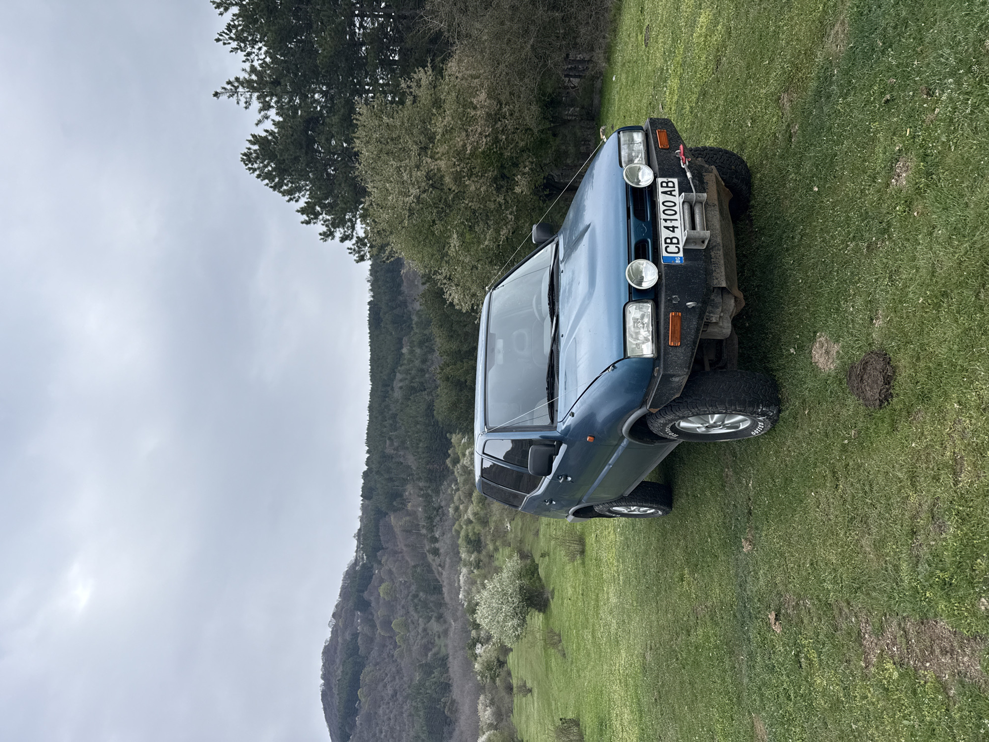 This image captures a robust blue or teal-colored SUV parked on a grassy slope in an outdoor, rural setting, identified as Buhovo, Bulgaria. The vehicle, possibly a Nissan Terrano or similar 4x4, faces towards the left (when the image is correctly oriented) and features modifications such as two prominent circular auxiliary lights and a winch mounted on its front bumper. Large, chunky all-terrain tires are visible on the wheels. A Bulgarian license plate, displaying "CB 4100 AB," is clearly visible on the front of the vehicle.
The foreground and midground consist of vibrant green grass, with some areas showing patches of drier earth or disturbed ground. In the background, a series of hills or mountains stretch across the horizon, densely covered with a mix of trees. Many deciduous trees show signs of early spring, with fresh green leaves or white blossoms, suggesting the photo was taken during this season. The sky is uniformly overcast and light grey, indicating a cloudy daytime. No people are visible, and the scene conveys a sense of tranquility in a natural, elevated landscape.