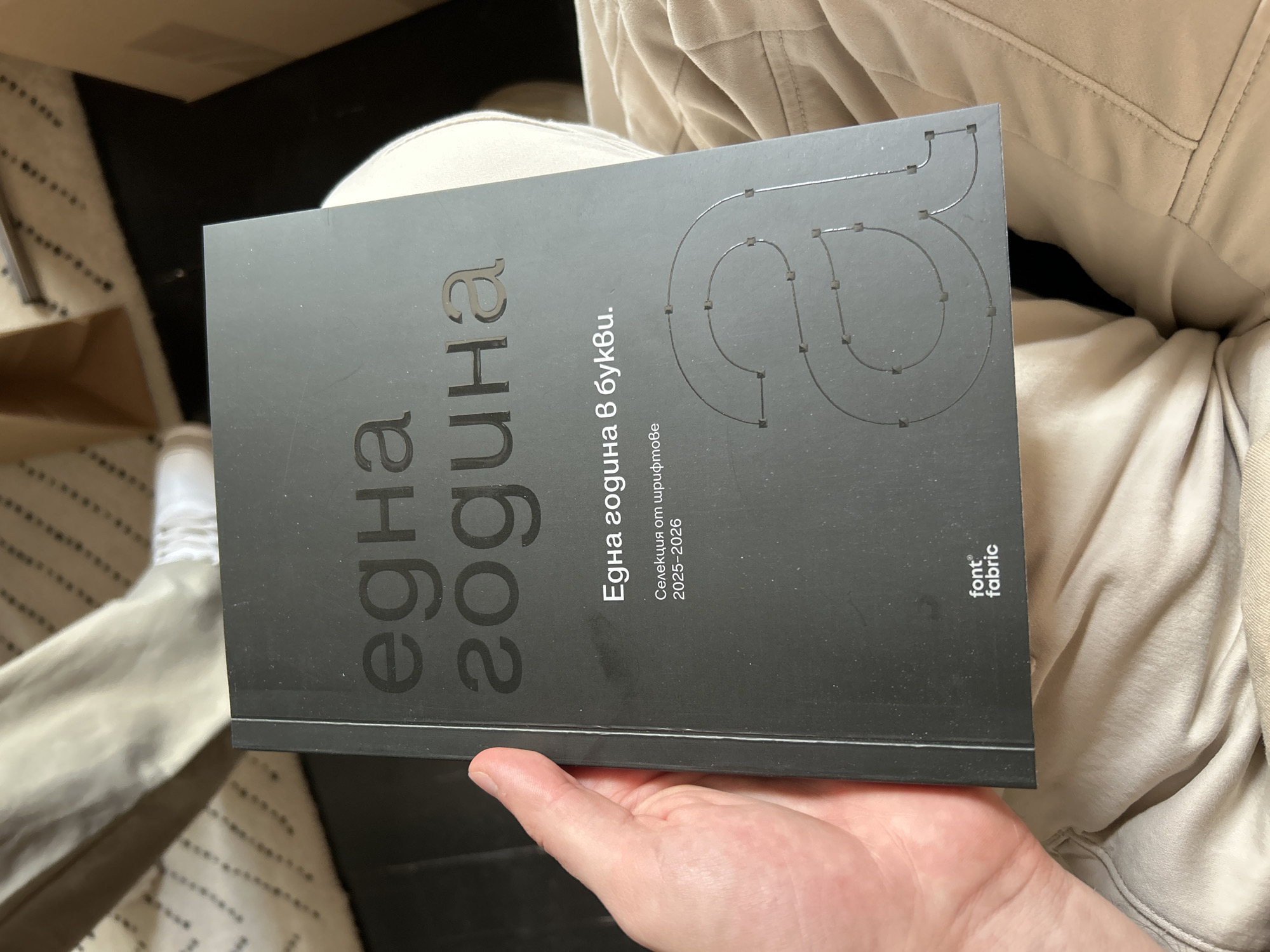 A hand holds a book in a casual setting. The book's cover is dark, featuring text in Cyrillic ("една година 8 букви" - "one year 8 letters"). The text "Селекция от шрифтове 2025-2026" ("Selection of Fonts 2025-2026") is also visible. The "font fabric" logo is located in the bottom-right corner. The subject is sitting on what appears to be a couch and is wearing light colored clothing. There is also a shoe on the floor nearby. It is not possible to infer the location (Sofia, Bulgaria) from the information available in the image.