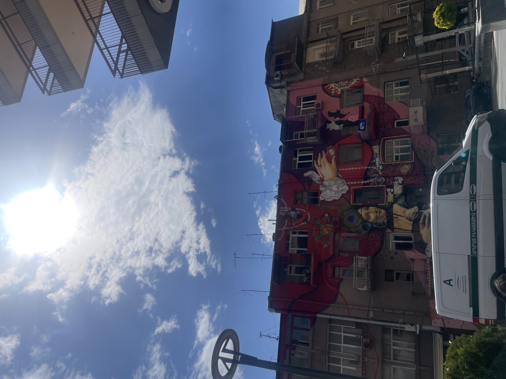 Captured from a low-angle perspective, the image showcases the vibrant architecture of Yerevan, Armenia. The sky is a clear blue, punctuated by scattered white clouds and a bright sun. A portion of a modern building with metal balcony railings is visible in the upper left.
The focal point is a tall building adorned with a large, colorful mural covering its exterior. The mural features a complex design with human figures, patterns, and bright hues of red, purple, and orange. Windows and balconies of the building are integrated into the artwork.
On the right side of the image, a white van is partially visible, obscuring a portion of the building. The van has a green stripe and some text in Armenian. A street lamp pole with a circular fixture at the top stands in the foreground.