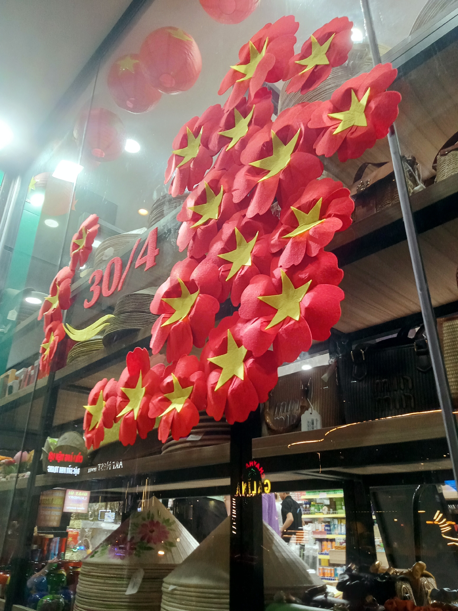 This image captures a reflection-filled view through a shop window, adorned with festive decorations in Nha Trang, Vietnam. The main subjects are numerous red floral shapes, each featuring a prominent yellow five-pointed star in the center, mimicking the design elements of the Vietnamese flag. These decorations are arranged dynamically across the upper portion of the window. Several round red lanterns, also with yellow stars, hang in the background, further enhancing the celebratory theme.
Prominently displayed in red numbers on the glass is "30/4", referring to April 30th, Vietnam's Reunification Day, indicating that the decorations are in observance of this national holiday.
Inside the shop, traditional Vietnamese conical hats (Nón Lá), some with painted floral designs, are stacked on shelves. Other shelves hold various items like woven baskets, bags, and what appears to be souvenirs or general merchandise. Reflections on the glass provide glimpses of the shop's interior, including a partially visible person (a man in a black shirt) and shelves stocked with products, possibly groceries or beverages, in the background. Bright interior lighting creates glare on the glass surface.
Visible text, often reflected or upside down, includes "30/4", and fragments like "VUI TẾT ĐOÀN..." (likely part of a holiday greeting), "HANDMADE" on a label, and other indistinct signage such as "...YANA" and "AIR THUY AN" (upside down). The scene depicts a festive atmosphere, likely a business decorated for the national holiday, with goods for sale to customers or tourists.