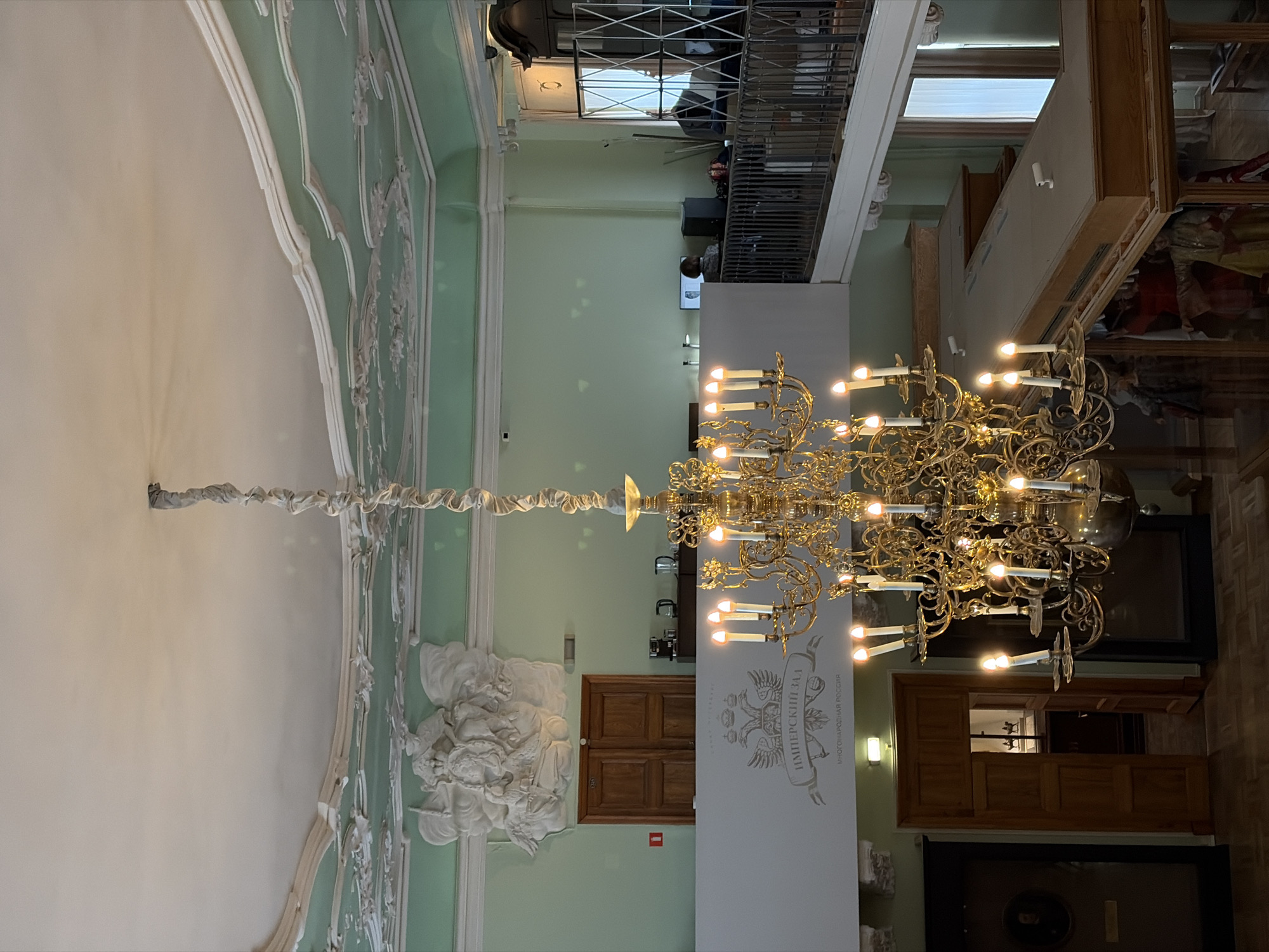 The image captures a grand interior space in Saint Petersburg, Russia. A large, ornate gold chandelier hangs prominently in the center, suspended from a wrapped cord extending upwards. The room features light green walls adorned with elaborate white plasterwork and decorations.
A balcony is visible in the upper portion of the image, with people walking around on it.
On one wall, there's a sculptural relief of figures, and a wooden door is positioned on the same wall. Adjacent to the door is a tall, white section of the wall bearing an inscription, possibly related to the "IMPERIAL HALL".
The overall environment appears to be within a historical building or museum.