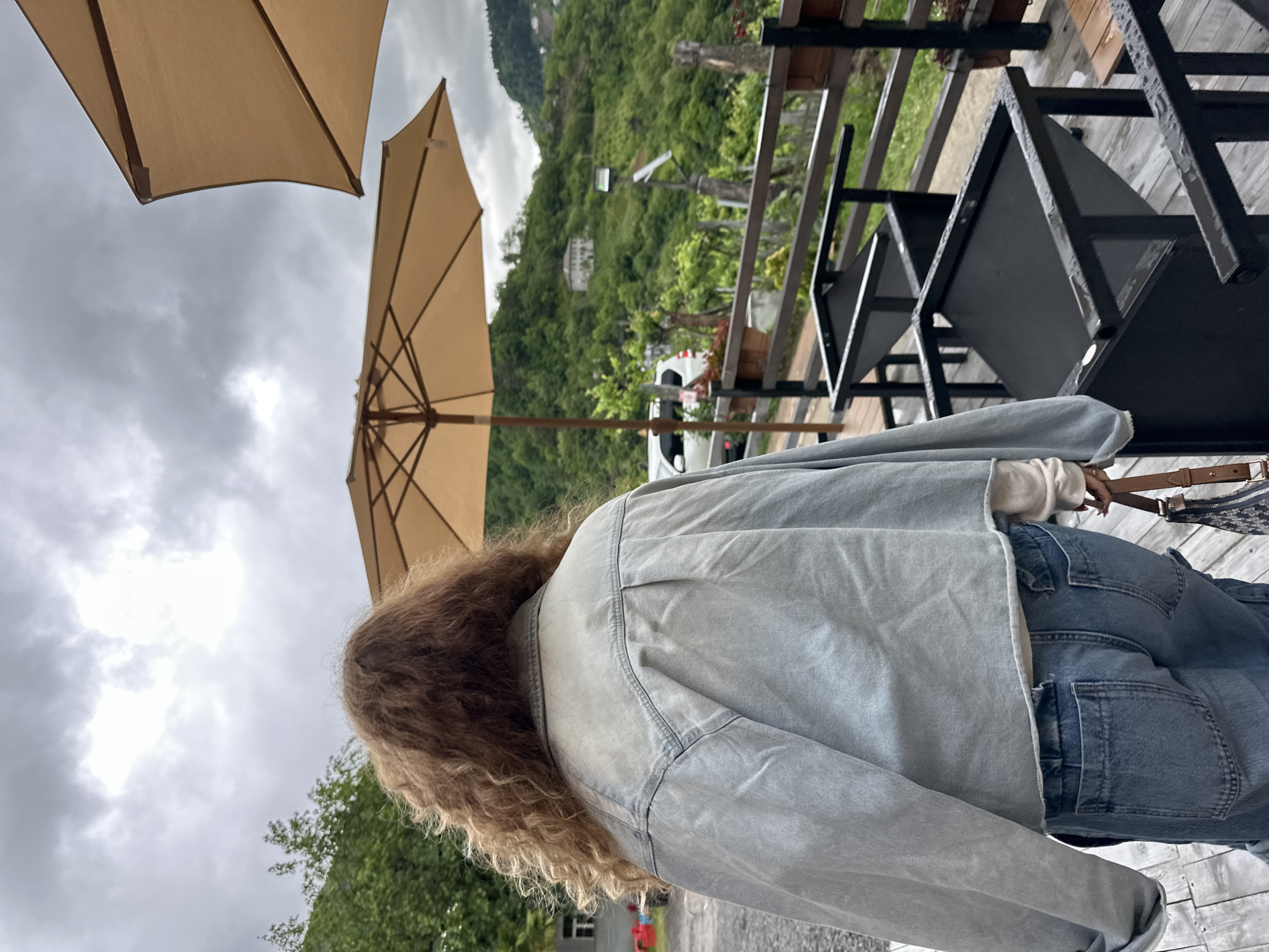 The image depicts a person from the rear, standing on a wooden deck. The person has curly, light brown hair and is wearing a light blue denim jacket and blue jeans. They are holding a brown and navy patterned handbag.
Two tan-colored patio umbrellas are visible, partially obscuring the overcast sky. In the background, there is a lush green hillside with some buildings visible. A black metal table and chair set are on the deck near the person.
The image is likely taken outdoors in a setting overlooking a natural landscape. The city of Tibeta, Georgia is not apparent from the given image.