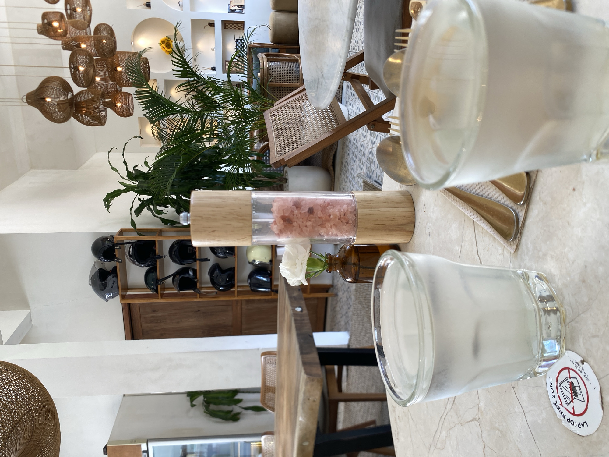 The image shows a bright, airy interior space that appears to be a cafe or restaurant. In the foreground, two glasses of water sit on a marble-patterned table. A wooden salt grinder filled with pink salt is positioned between the glasses. A single white flower in a brown glass vase adds a touch of decor. Behind the table, a wooden chair with a woven seat is visible, as well as a larger wooden table. Further back, a shelf unit displays several black helmets. Lush green plants are scattered throughout the space, contributing to a natural, relaxed atmosphere. Woven pendant lights hang from the ceiling. The overall impression is one of a modern, stylish establishment. The text on the sticker in the lower left corner of the image indicates "LAPTOP FREE ZONE," suggesting this is a place intended for disconnection and relaxation. The presence of the sticker implies the location is likely a cafe or dining establishment in a tourist-oriented area. Considering the context of Bali, Indonesia, and the style of the cafe, it is plausible this establishment is located in the popular area of North Kuta, known for its trendy cafes and beaches.