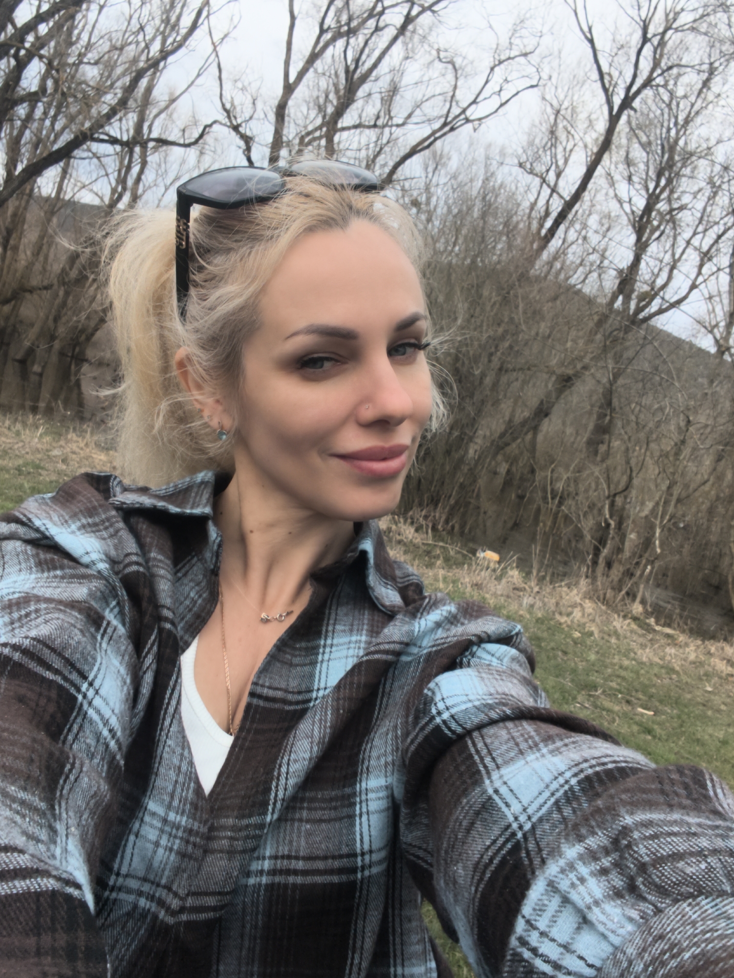 A blonde woman is taking a selfie in what appears to be a natural setting. She has sunglasses perched on top of her head and is wearing a blue and brown plaid shirt over a white top. She has fair skin and is wearing a necklace. The background consists of trees without leaves and some grassy areas. The overall impression is a casual outdoor setting. The location is near Орлиновский округ, Ukraine.