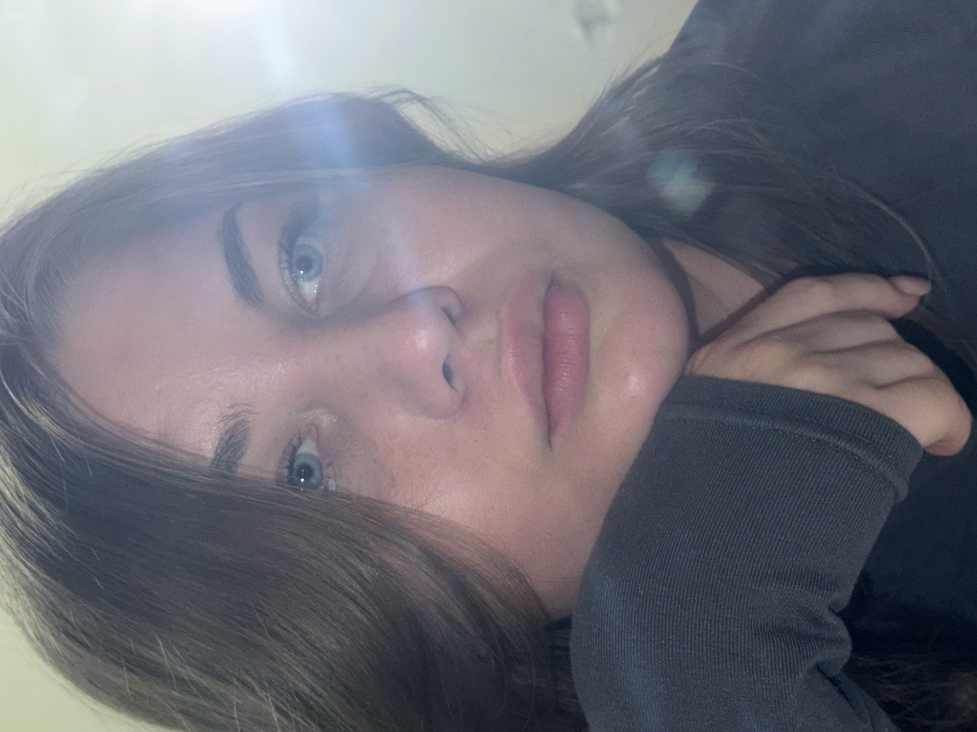 The image depicts a woman with light skin, blue eyes, and brown hair. She is wearing a dark grey long-sleeved shirt. Her face is in focus, and her expression appears neutral. She rests her head on her hand/sleeve. A bright light source illuminates the upper portion of her face, causing lens flare in the image. There is no visible information to determine that the image was taken in the city of Chaikovsky, Russia.