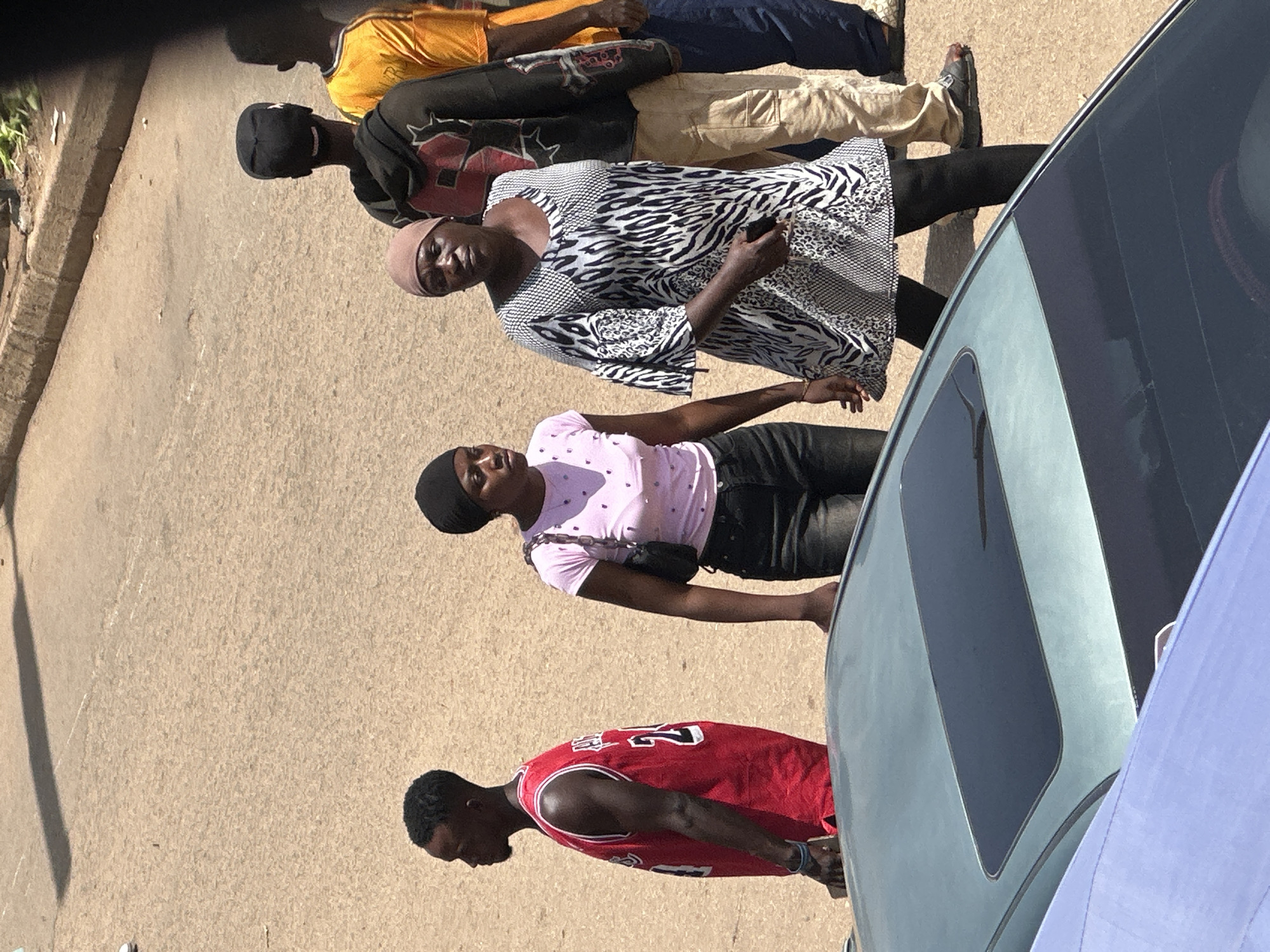 In Bukuru, Nigeria, a group of four young adults stands by a light blue car on a paved surface. The scene appears to be outdoors on a bright, sunny day.
From top to bottom, the individuals are:
A person wearing a yellow t-shirt and dark pants.
A person in a dark shirt with red and white accents on the chest, a black beanie, and tan pants.
A woman wearing a light pink t-shirt with a speckled pattern and black pants. She has a black crossbody bag.
A young man wearing a red basketball jersey with the number 23 on the back, black shorts, and black sneakers.
The people seem to be waiting or standing around, with no specific activity clearly depicted. The sun casts distinct shadows, suggesting it is likely midday. The setting appears to be a street or a parking area. The car's tinted windows are visible. The overall mood is casual. No visible text is discernable on any of the clothing or objects.