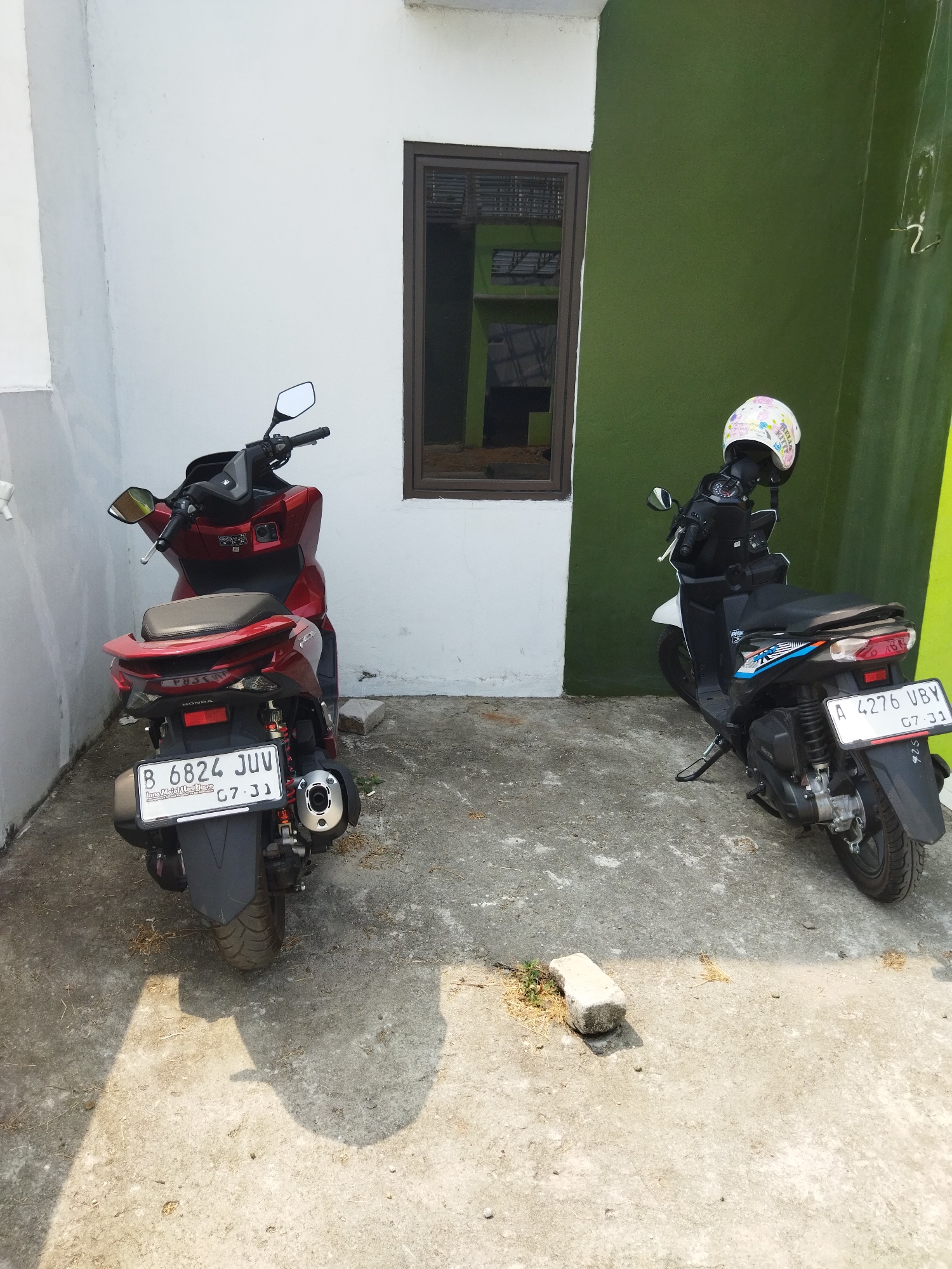 The image depicts an outdoor scene in the city of Suka Sari, Indonesia, featuring two parked scooters in front of a building. The building has a white wall on the left and a green wall on the right, with a dark-framed window visible on the white section. The ground is paved with concrete, showing some debris and shadows cast by the scooters and the building.
On the left, a red scooter is parked. It has a license plate that reads "B 6824 JUV". The scooter is facing away from the camera, and its rear end with taillights and exhaust pipe is clearly visible.
On the right, a white and black scooter is parked. It has a license plate that reads "A 4276 UBY". A white helmet with a pink Hello Kitty design is placed on the handlebars of this scooter. This scooter is also facing away from the camera.
The overall lighting suggests it is daytime, with bright sunlight creating distinct shadows on the ground. There are no people or animals visible in the image. The environment appears to be a residential or commercial area with a simple, functional architectural style.