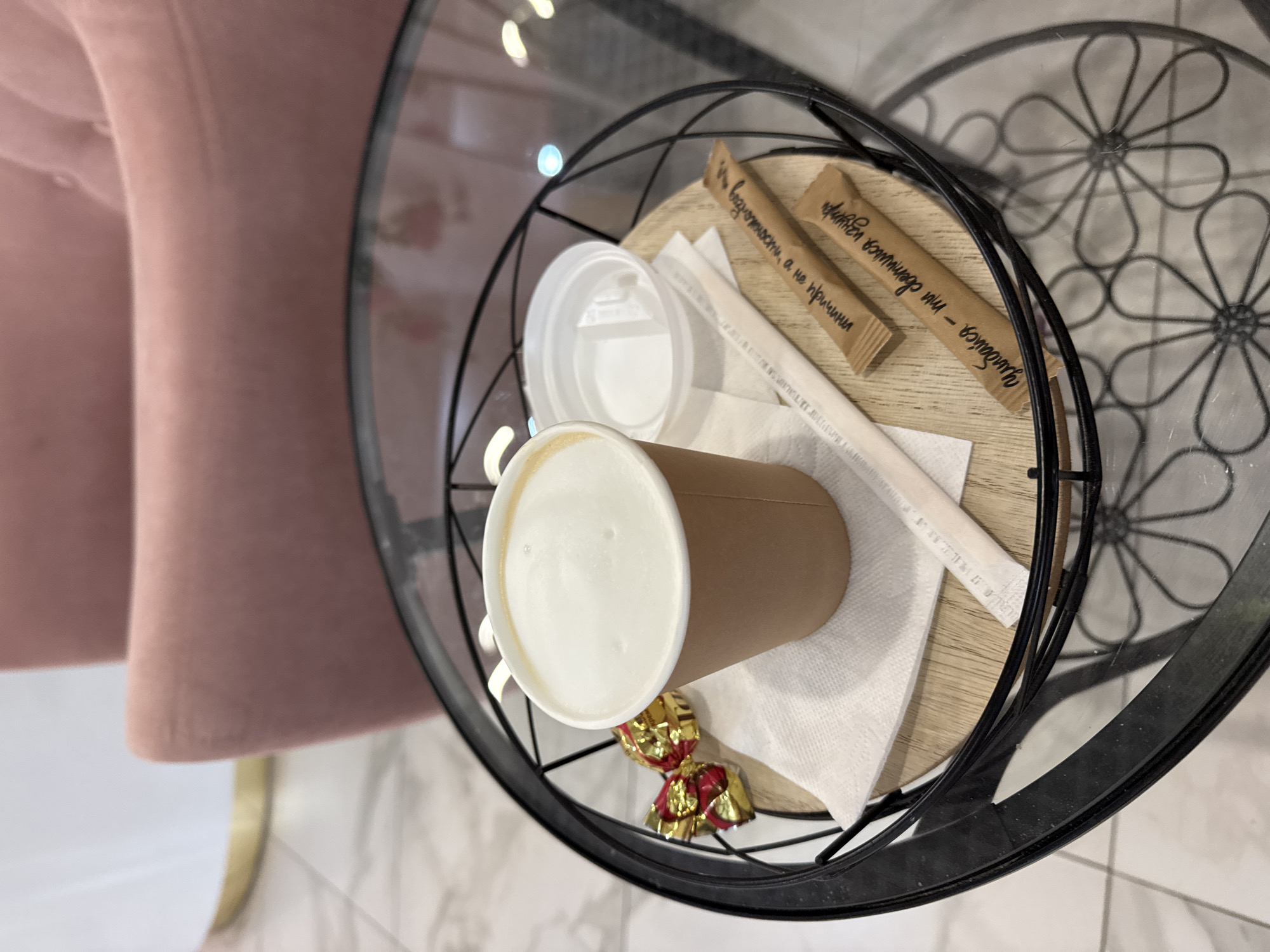 A coffee cup with a lid, a foil-wrapped chocolate, and two sugar packets are arranged on a wooden board within a black wire basket on a glass-top table. The basket is placed on a tiled floor in front of a plush pink armchair and a tall white cylindrical object. The coffee cup is made of brown paper and contains a frothy beverage. One sugar packet has the inscription "Улыбайся - ты светишься изнутри" and the other has "а не кричи". A white straw is also present on the board. The environment suggests an indoor cafe or lounge setting.