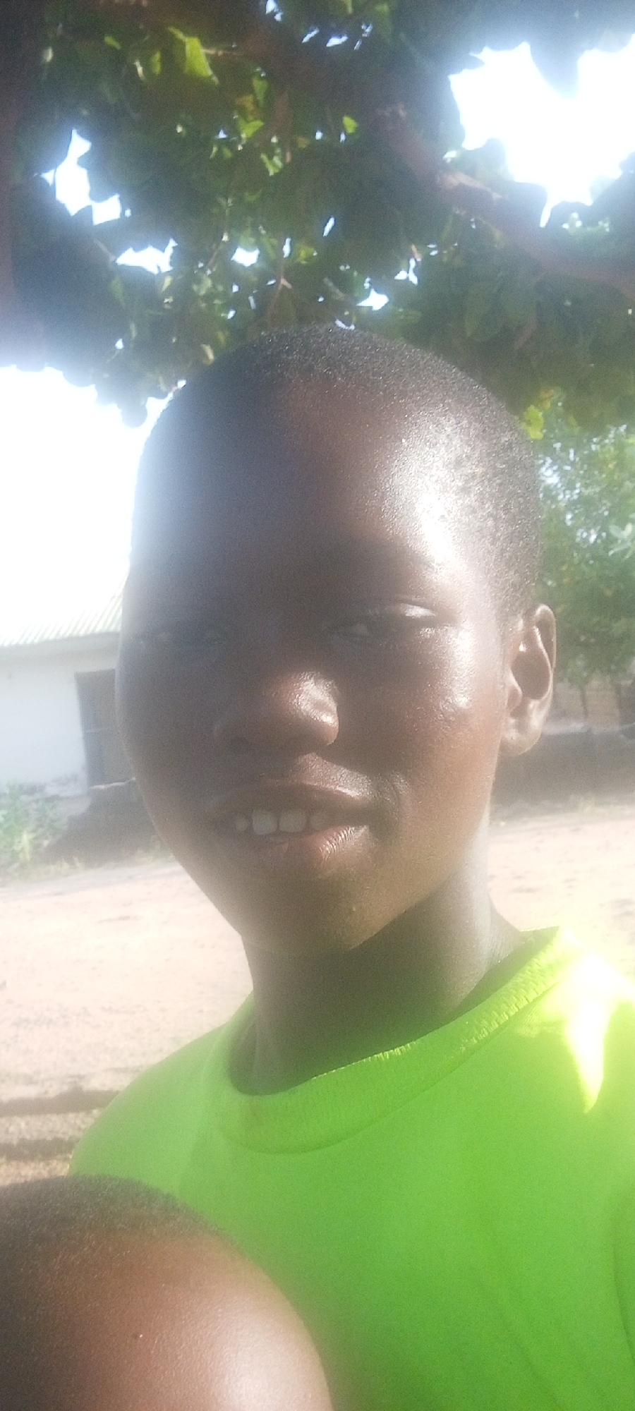 A close-up shot of a young person with dark skin and short hair, wearing a bright green t-shirt. The person is smiling slightly, showing their teeth. The background shows a building with a window and a sandy area, suggesting an outdoor location. There are also branches and leaves overhead, indicating a possible tree or vegetation cover. The light is bright, creating some glare on the subject's face. The location is potentially Adankolo, Nigeria, based on user prompt.