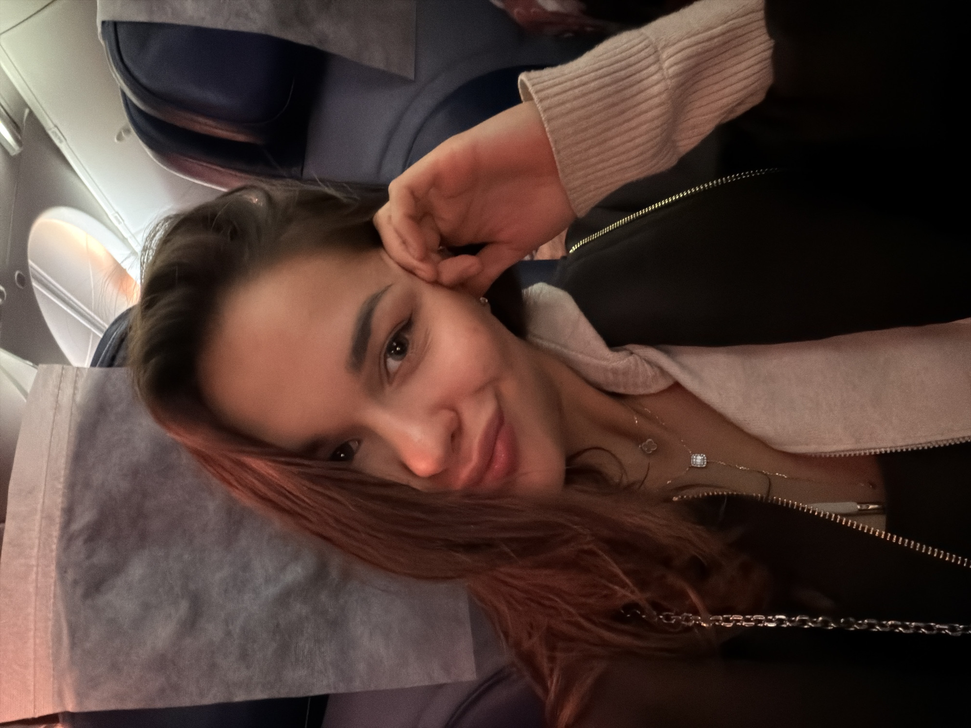 A woman is reclining on an airplane seat. She has medium-length reddish-brown hair, and she is wearing a black jacket over a light-colored sweater. She is looking at the camera and smiling slightly, with one hand near her ear. The airplane window and seat are visible in the background, as is a grey headrest cover. There is no information to suggest that this image was taken in Sochi, Russia.