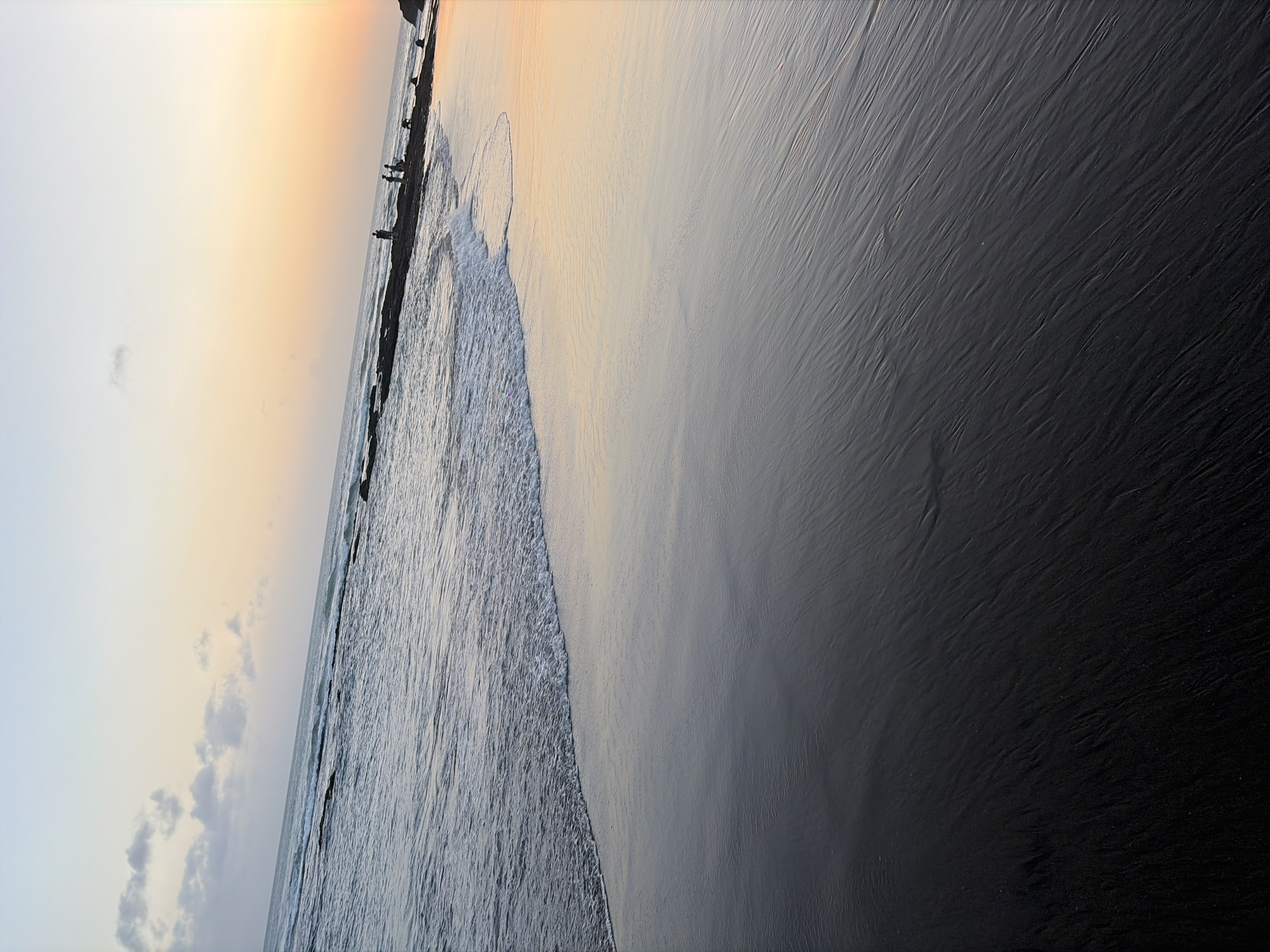 The image depicts a coastal scene at sunset in North Kuta, Indonesia. The foreground features dark, wet sand with visible ripple patterns created by receding waves. The midground shows a gentle wave breaking onto the shore, with the wet sand reflecting the warm hues of the sunset. In the background, a line of dark rocks juts out into the ocean, and several silhouetted figures of people are visible standing on these rocks. The sky exhibits a gradient of colors, transitioning from a pale blue at the top to soft orange and pink near the horizon, indicative of a sunset. The ocean itself appears calm with small waves.