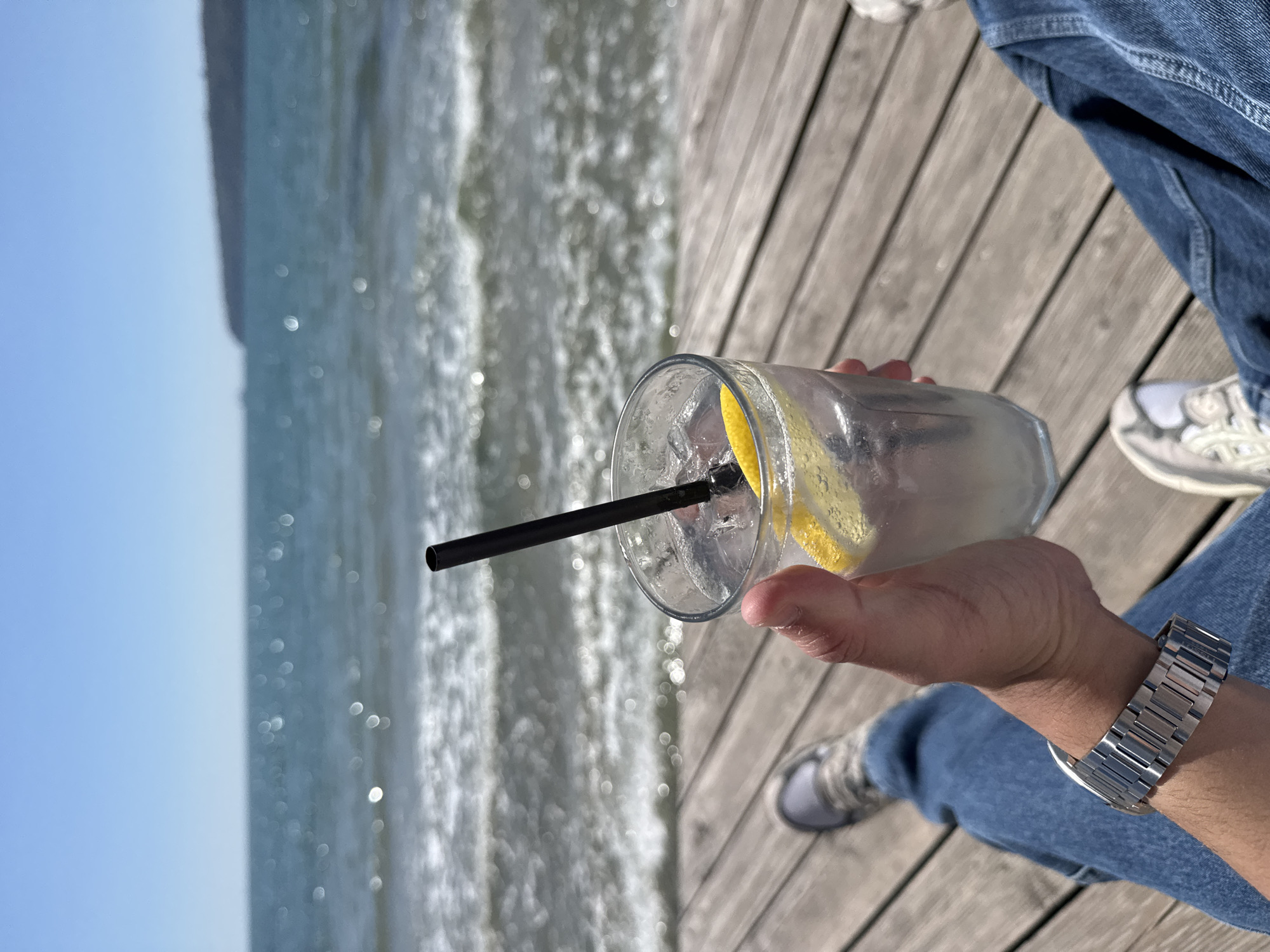 The image depicts a person holding a glass of lemonade with a black straw and lemon slices, sitting on a wooden pier. The background shows the sea and clear sky. The person is wearing blue jeans and a silver wristwatch. Footwear consists of two distinct sneakers. The location is likely the beach area in Varna, Bulgaria, suggested by the presence of the sea and pier-like structures commonly found in coastal areas.