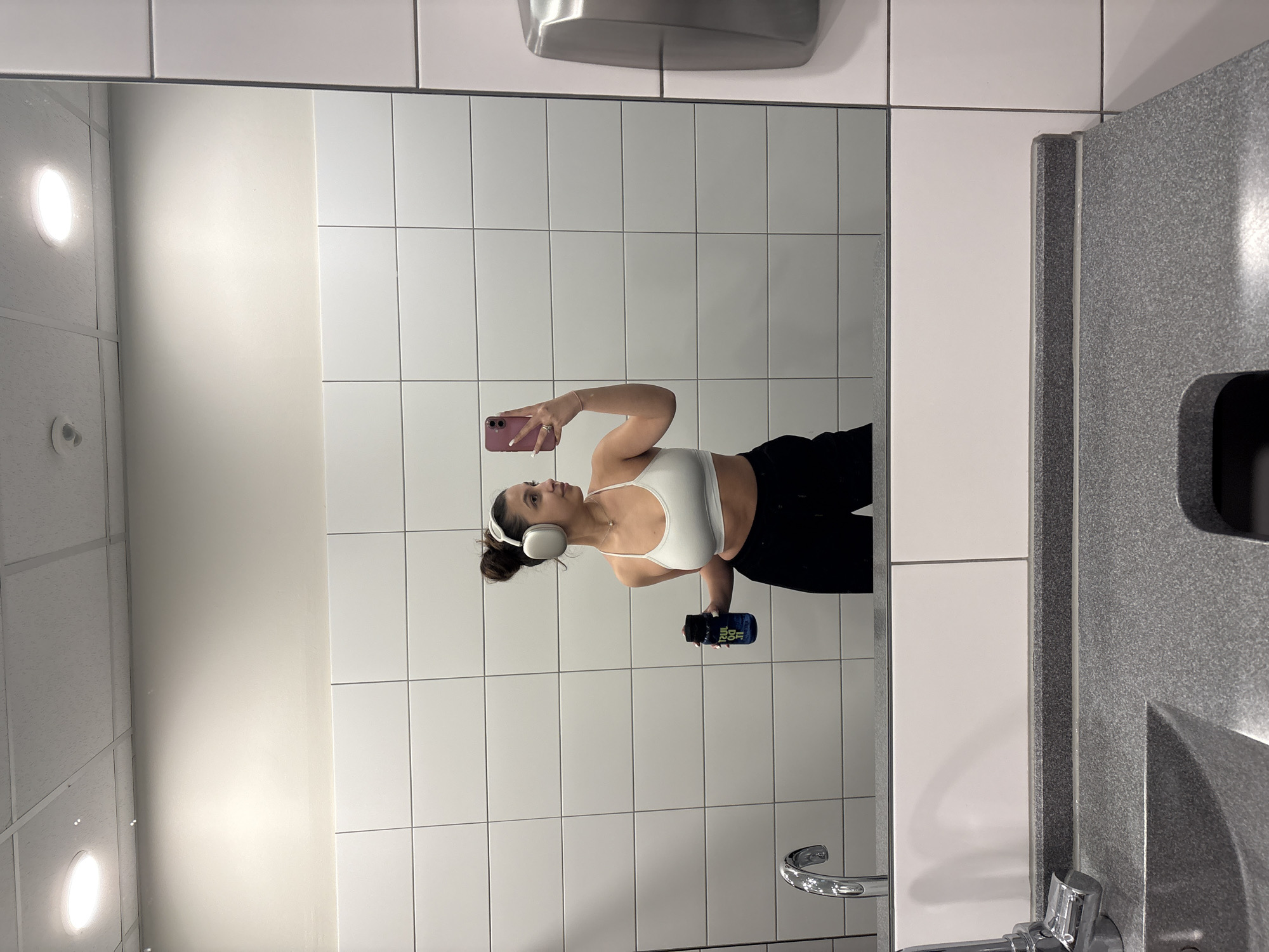 A young woman is taking a mirror selfie in a tiled bathroom. She is wearing a white sports bra, black pants, and white headphones. She is holding a water bottle in one hand and a pink smartphone in the other. The bathroom has white square tiles on the walls, a white ceiling with recessed lighting, a stainless steel hand dryer, and a gray countertop with a sink and faucet. The location is in Mönchengladbach, Germany.