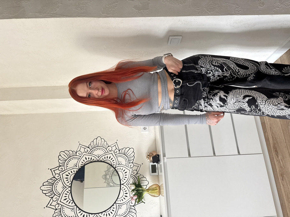 An image taken indoors in what appears to be a residential setting in Rostov-on-Don, Russia, depicts a female subject standing centered, looking directly at the camera with a subtle smile. She possesses long, straight, vibrant red hair parted in the middle and fair skin with visible freckles on her nose and cheeks. She is attired in a light grey long-sleeved crop top featuring a wrap-around midriff design and black wide-leg jeans prominently adorned with white dragon motifs. A black belt with silver eyelets and a heart-shaped silver buckle secures the jeans. A silver-toned smartwatch is present on her left wrist, and a tattoo with an abstract design is visible on her left forearm. Her left hand is casually placed in her jeans pocket.
The background consists of textured white walls and a light wood laminate floor. On the left wall, a round mirror with a black frame is centrally mounted, surrounded by a large, intricate black mandala-style wall decal. Beneath the mirror, a white dresser or cabinet with multiple drawers/doors holds several items: a golden fluted vase containing pink tulips, a digital clock, and a small decorative hedgehog figurine holding a dark-colored smartphone in a stand. A white electrical outlet is visible on the wall beneath the mirror, and another white electrical fixture is present on the right wall, which features a vertical structural element or seam. The overall lighting suggests an interior, possibly daylight-illuminated, scene.