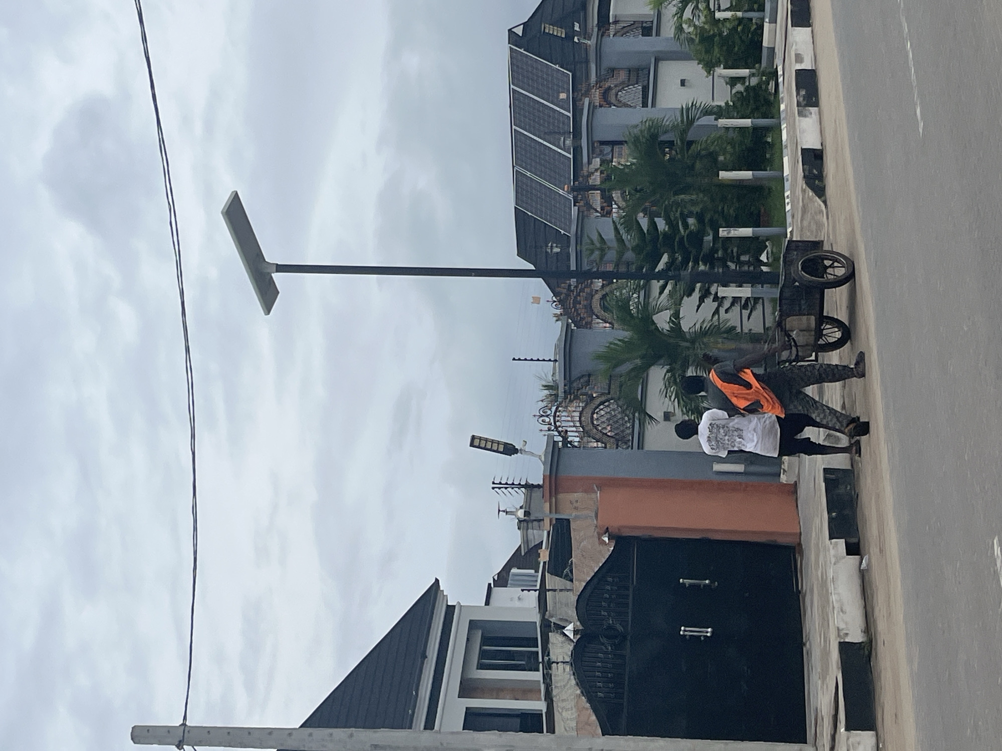 Two men are walking along a street in Badagry, Nigeria, pushing a four-wheeled cart containing unspecified items. One man wears an orange safety vest over dark clothing, while the other wears a white t-shirt and dark pants. The street runs between residential buildings, including a two-story building with solar panels on the roof and another with a black gate. A modern solar street lamp is visible, along with overhead electrical wires. The sky is overcast. The road is paved and has a painted curb.