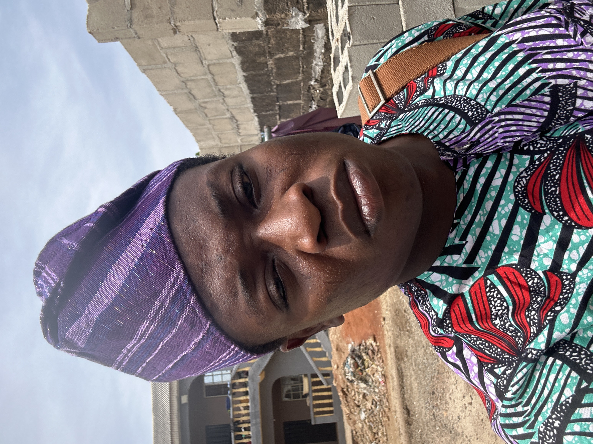 A close-up, angled portrait of a person with dark skin wearing a purple striped head covering and a brightly patterned shirt with colors including purple, green, red, white, and black. The person's eyes are closed or nearly closed. Behind the person, there is a concrete block wall. The location is Unknown, Unknown.