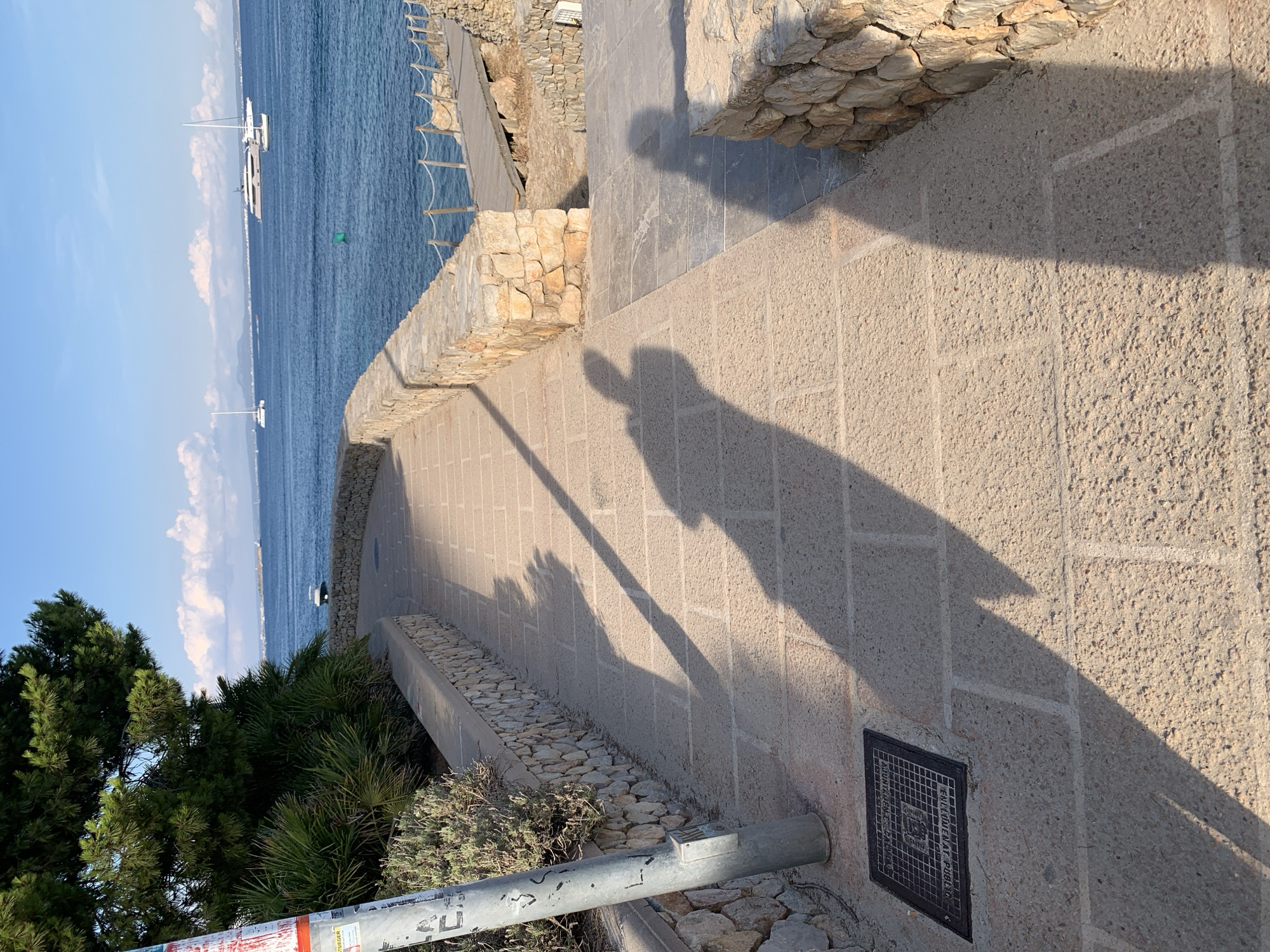 The media file depicts a sun-drenched scene in Calvià, Spain. The foreground is dominated by a textured, light-colored pavement, likely stone tiles, stretching diagonally across the frame. A prominent shadow, cast by a person holding a camera, stretches across the pavement, indicating it's late afternoon or early evening as the sun is low in the sky. The shadow shows the outline of a person with their arm raised, suggesting they are taking the photograph.
To the left, a stone retaining wall borders a steep incline covered in lush green trees and shrubs. Beyond this, the deep blue sea stretches to the horizon under a bright blue sky dotted with fluffy white clouds. Several white sailboats are visible on the water, one closer and larger, another further out. A small green buoy floats in the sea.
The background features a built stone wall, characteristic of Mediterranean architecture, and a pathway leading further up. The overall impression is one of a beautiful coastal location on a clear, sunny day. The scene evokes a sense of leisure and vacation. There is no visible text in the media file.
