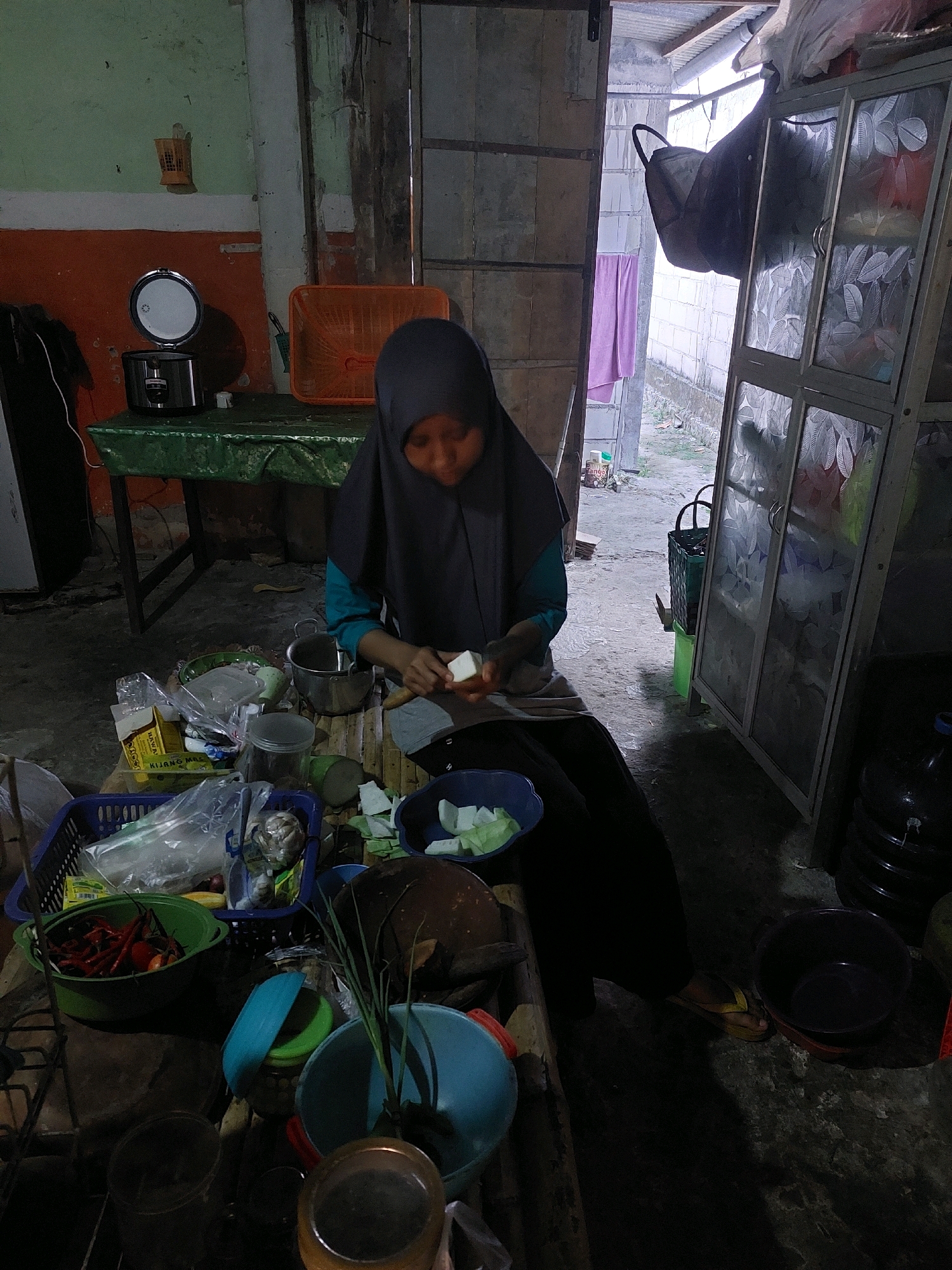 In SARANGAN, Indonesia, an individual wearing a dark hijab and teal shirt is seated on the floor of a rustic kitchen, preparing food. The person is in the process of slicing a white food item, likely a vegetable or tofu, with a knife held in their right hand. A small, light-colored piece of the food item is held in their left hand.
The environment appears to be a well-used, somewhat cluttered space. To the left, an orange wall meets a pale green painted section. A rice cooker sits on a green-covered table in the background. In front of the individual, a collection of kitchen items is spread out on a low wooden surface. These include various bowls, containers, plastic bags filled with ingredients, a green bowl overflowing with red chilies, and a blue bowl containing sliced cucumbers. A large stainless steel pot is positioned behind the person.
The background reveals an open doorway leading to an outdoor area with a white brick wall and a corrugated metal roof. A large, semi-transparent cabinet with decorative glass doors stands to the right, and a large black water jug is visible next to it. The floor is a rough, concrete surface. The lighting is dim, with shadows cast throughout the scene, suggesting it is either early morning or late evening.