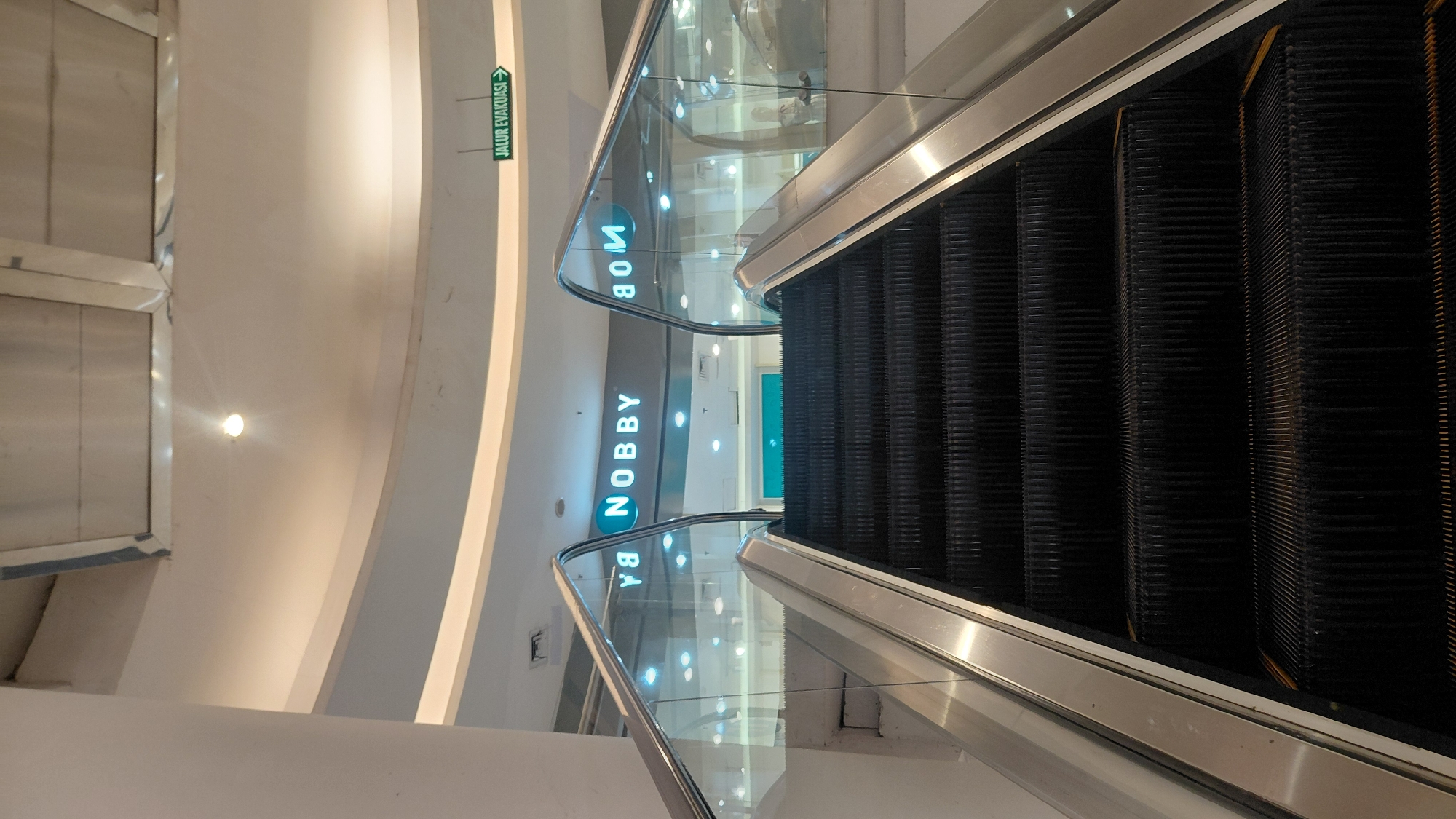The image shows an upward-facing shot of an escalator in a shopping mall. The escalator has black treads with yellow accents on the edges. The handrails are made of polished metal and are supported by glass panels. In the background, a sign with blue lettering on a grey background reads "NOBBY". To the right, a green sign with white text and an arrow indicates "JALUR EVAKUASI" (evacuation route). The ceiling above is illuminated by soft lighting, with a curved white structure. The overall environment appears to be clean and modern, typical of an indoor shopping center. The location is likely a mall in Purwokerto, Indonesia, as suggested by the Indonesian text on the evacuation sign. There are no people visible in the image.
