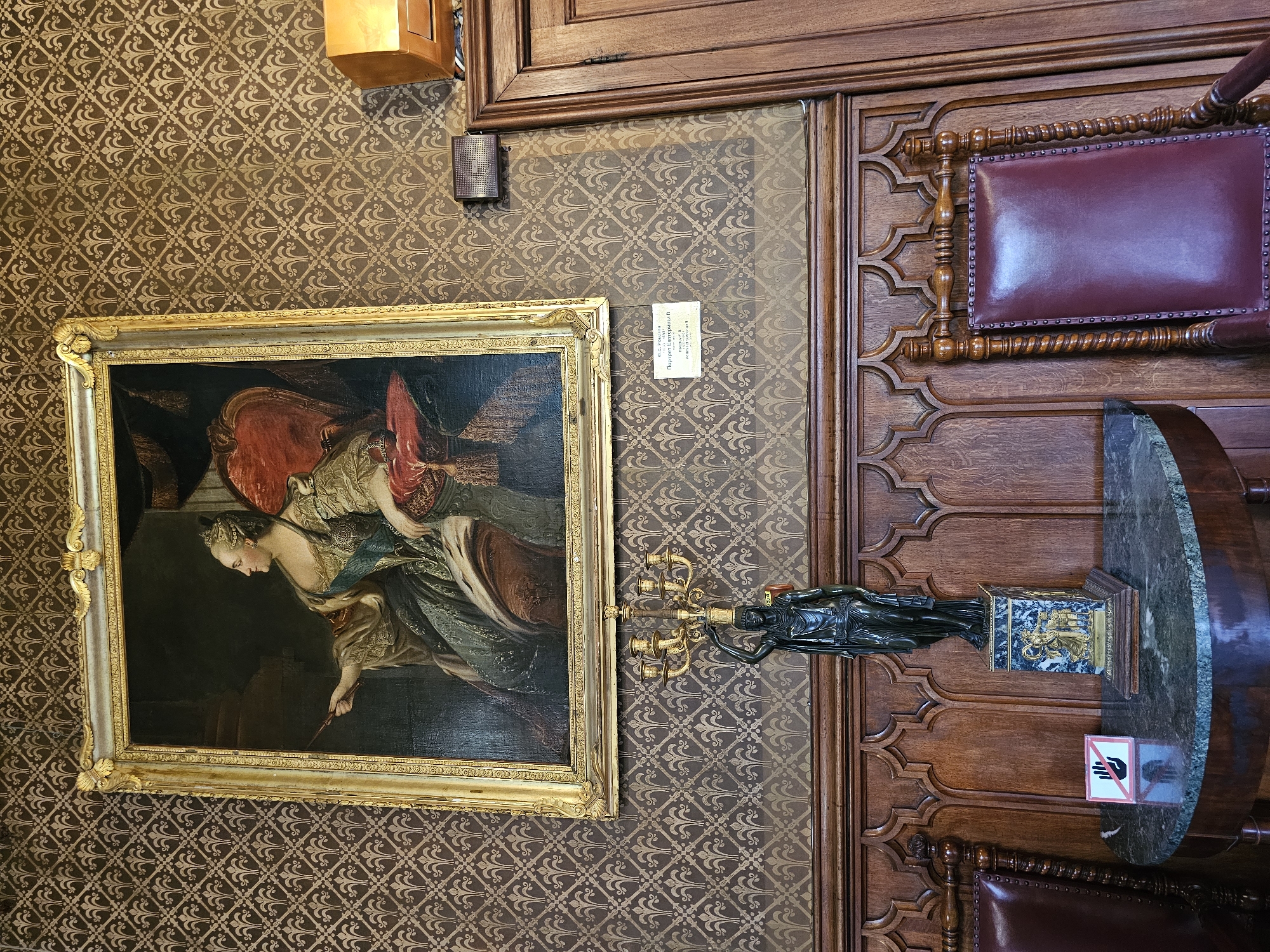A photograph shows a framed painting hanging on a patterned wall, alongside a decorative wooden panel and a statue on a marble-topped table, likely taken in a historical or cultural institution.
The painting, set in a gold frame, depicts a woman in a historical dress. To the right of the painting, a bronze statue of a woman holding a candelabra is placed on a pedestal on a table with a marble top. A "Do Not Touch" sign is visible on the table. The wooden panel behind the table features ornate carvings and a leather-upholstered seat.
The wallpaper has a repeating decorative pattern in shades of brown and gold. A small electrical box is visible above the painting. The location is possibly in Алупкинский городской совет, Ukraine, based on the file name metadata.