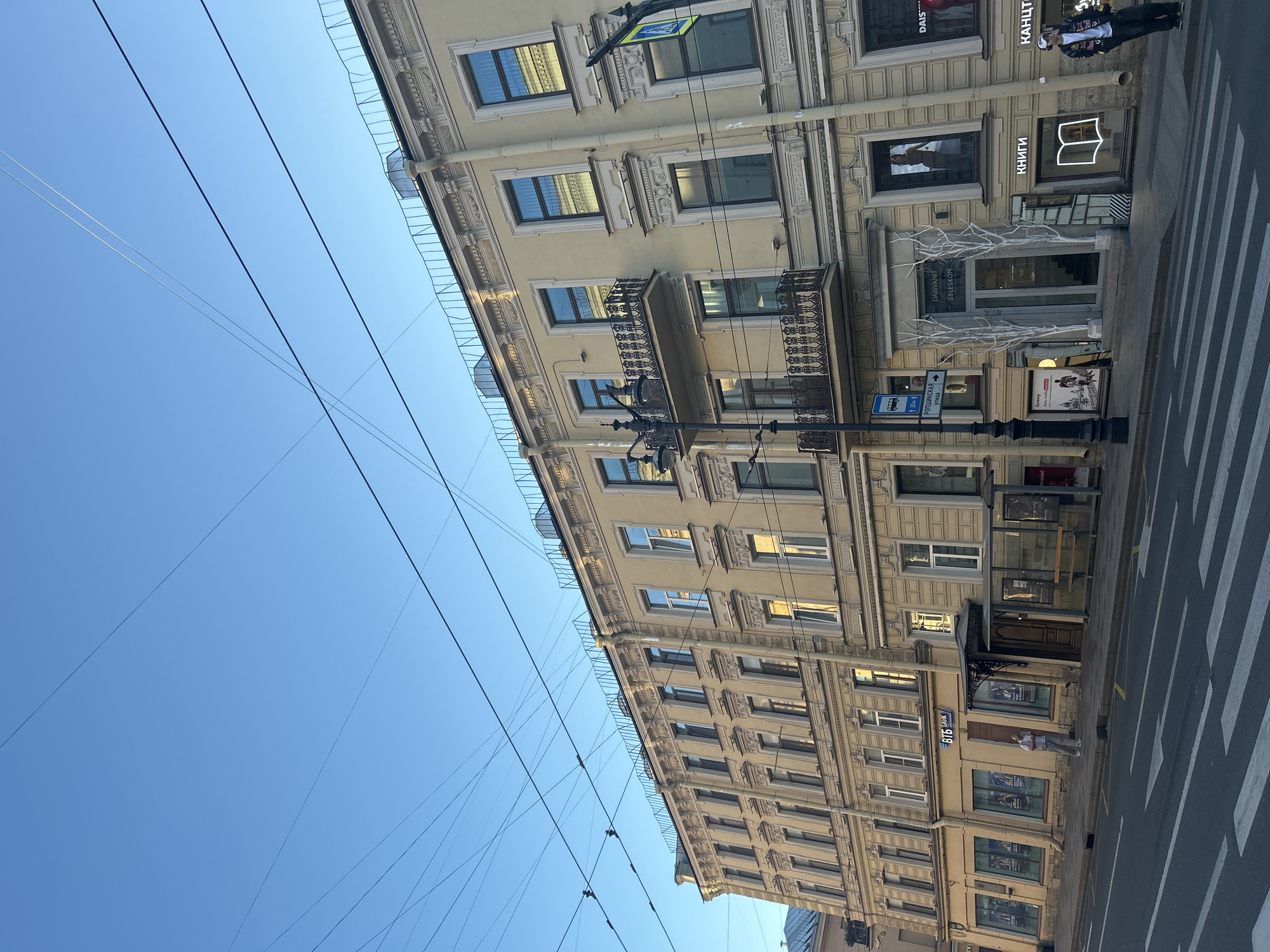 This image depicts a street scene in an urban environment, likely in Eastern Europe given the Cyrillic signage on the buildings. A multi-story building with classical architectural features, painted in a pale yellow hue, dominates the frame. The building has numerous windows, some with ornate balconies, and is adorned with decorative moldings.
Several businesses are visible at street level. One storefront displays the logo "ВТБ БАНК". Another appears to be a clothing store called "DAISYKOM". A bookstore with an illuminated open book sign is also present. The building is equipped with overhead power lines, characteristic of older city infrastructure.
A crosswalk is visible in the foreground, indicating a pedestrian crossing. A bus stop shelter with a bench is situated on the sidewalk. A few individuals are present: one man stands near the bank entrance, and another person, dressed in a baseball cap and jacket, stands near the bookstore. The sky above is a clear, light blue, suggesting daytime. The lighting indicates it is either early morning or late afternoon. The overall impression is that of a typical city street with commercial activity.