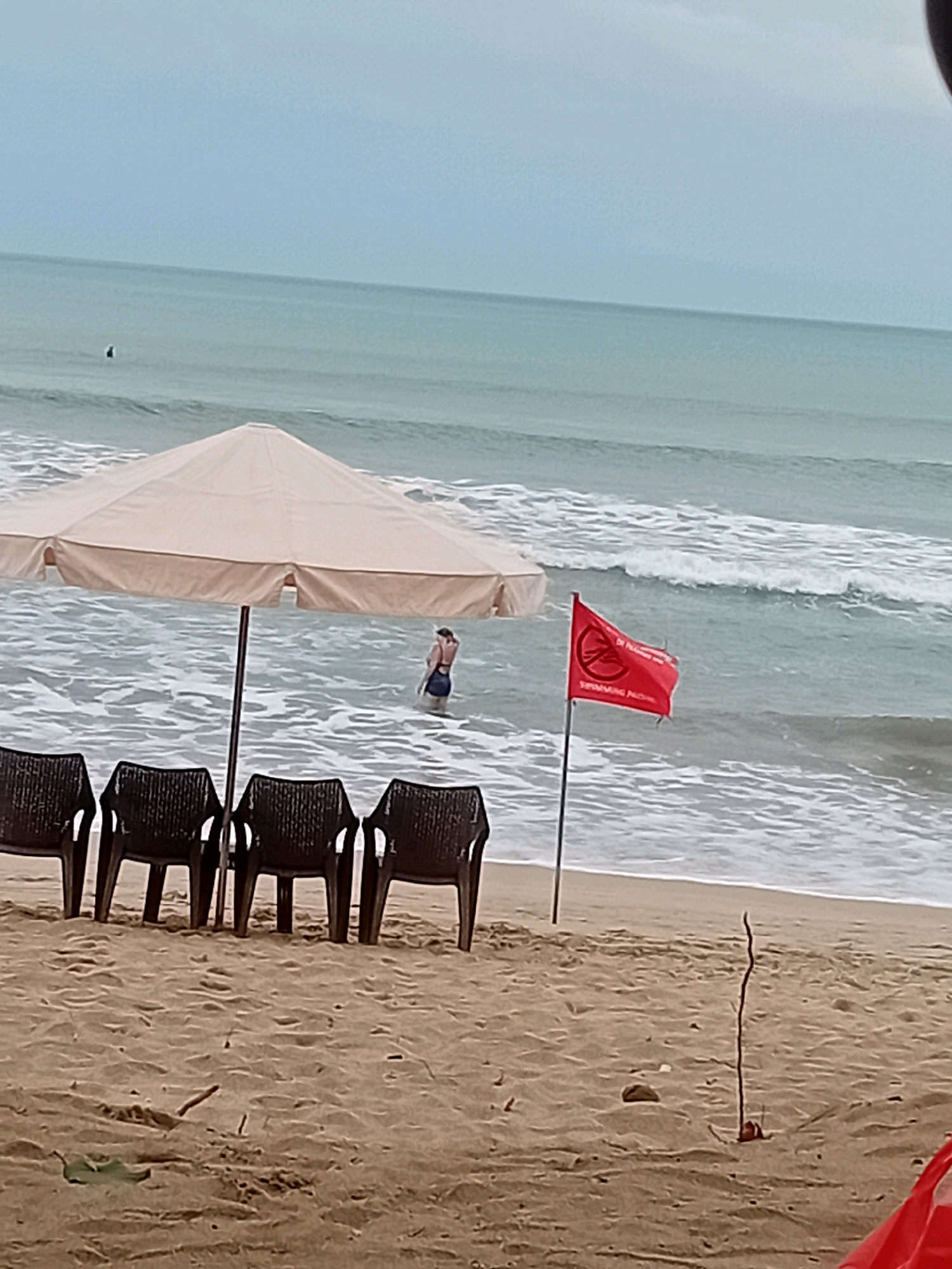The image shows a beach scene in Kuta, Indonesia. In the foreground, there are four empty dark brown plastic chairs arranged in a row on the sandy beach. Behind the chairs, a large, light-colored beach umbrella is set up, providing shade. In the shallow water, a person wearing dark swimwear is standing, partially submerged. A red flag with a white circular symbol and text is planted in the sand near the water's edge, indicating a warning. The ocean is calm with small waves breaking on the shore under an overcast sky.