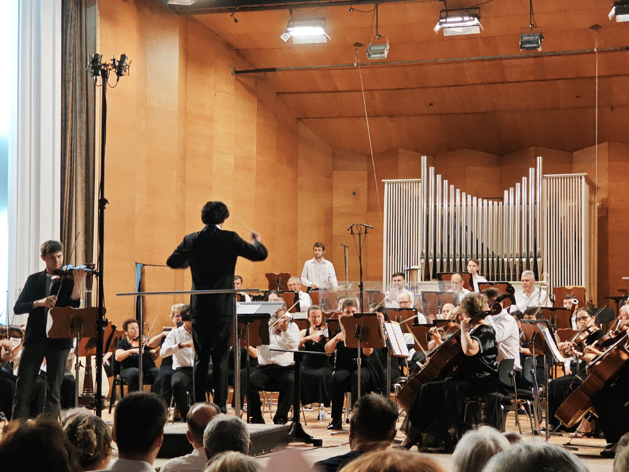 In an indoor concert hall with warm orange walls and wooden paneling, an orchestra performs on a raised stage. In the foreground, the back of a conductor in a black suit is visible, facing the orchestra and gesturing with a baton. To the left, a young man in a black jacket and dark trousers plays the violin. The orchestra comprises numerous musicians, many holding string instruments like violins and cellos, as well as wind instruments. Some musicians are seated, while others stand. Behind the orchestra, a large pipe organ with polished metal pipes dominates the background. Microphones on stands are positioned around the stage. The audience is visible in the lower portion of the frame, with their backs to the camera, suggesting the photograph was taken from within the audience. The lighting appears to be from overhead fixtures.