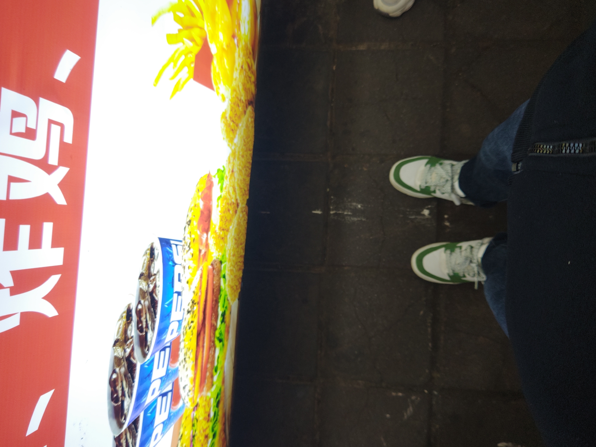 A ground-level shot shows a person wearing blue jeans and white sneakers with green accents, standing on a gray tile floor. A large advertisement is partially visible to the left, featuring images of Pepsi cans, a burger, and french fries, as well as red Chinese writing. The location is likely Shapingba District, China, given the presence of Chinese writing.