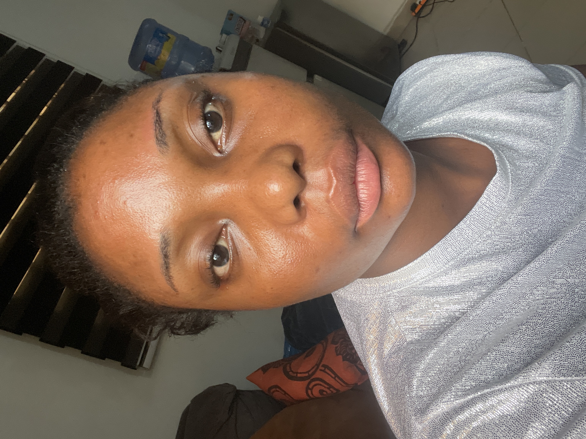 The image is a close-up, rotated portrait of a person, likely a woman, captured indoors in Abuja, Nigeria. The subject has dark skin and dark, curly hair that appears pulled back. She looks directly into the camera with a neutral expression, her eyes open and lips full and glossy.
She is wearing a silver, shimmery top, the fabric of which has a textured, reflective quality, appearing to sparkle with small, bright dots. Her skin shows natural texture and a healthy glow.
The background is dimly lit and somewhat cluttered. To the upper left and behind her head, dark horizontal blinds or louvers are visible. Above and slightly behind her, a blue plastic water bottle with an unreadable label can be seen. Below her head and to the right, an orange pillow with a dark, abstract swirling pattern is partially visible. The lighting is soft and somewhat diffused, suggesting an indoor setting with either low natural light or artificial illumination. The shot has a candid, personal feel.