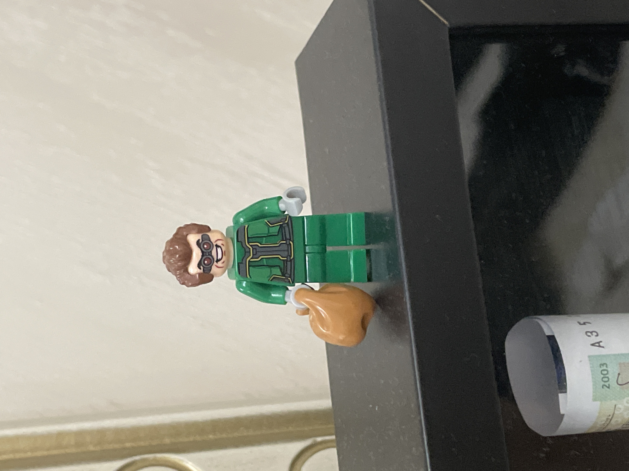 A Lego minifigure, resembling Dr. Octopus with a green suit, brown hair, and goggles, stands upright on a dark, flat surface. The minifigure's face features a wide, grinning expression. Its torso is decorated with black and yellow lines, suggesting a suit or uniform. In its right hand, the minifigure holds a light brown, shaped accessory that appears to be a Lego turkey leg or ham.
The dark surface beneath the minifigure extends to the right, forming what looks like the top edge of a frame or a shelf. To the far right, a rolled-up piece of white paper or currency rests on this dark surface. Visible text on the rolled paper includes the numbers "2003" and the characters "A35". The background is a plain, light-colored wall, indicating an indoor setting. The scene is static, showcasing the minifigure as a display. This setting is located in Ihtiman, Bulgaria.