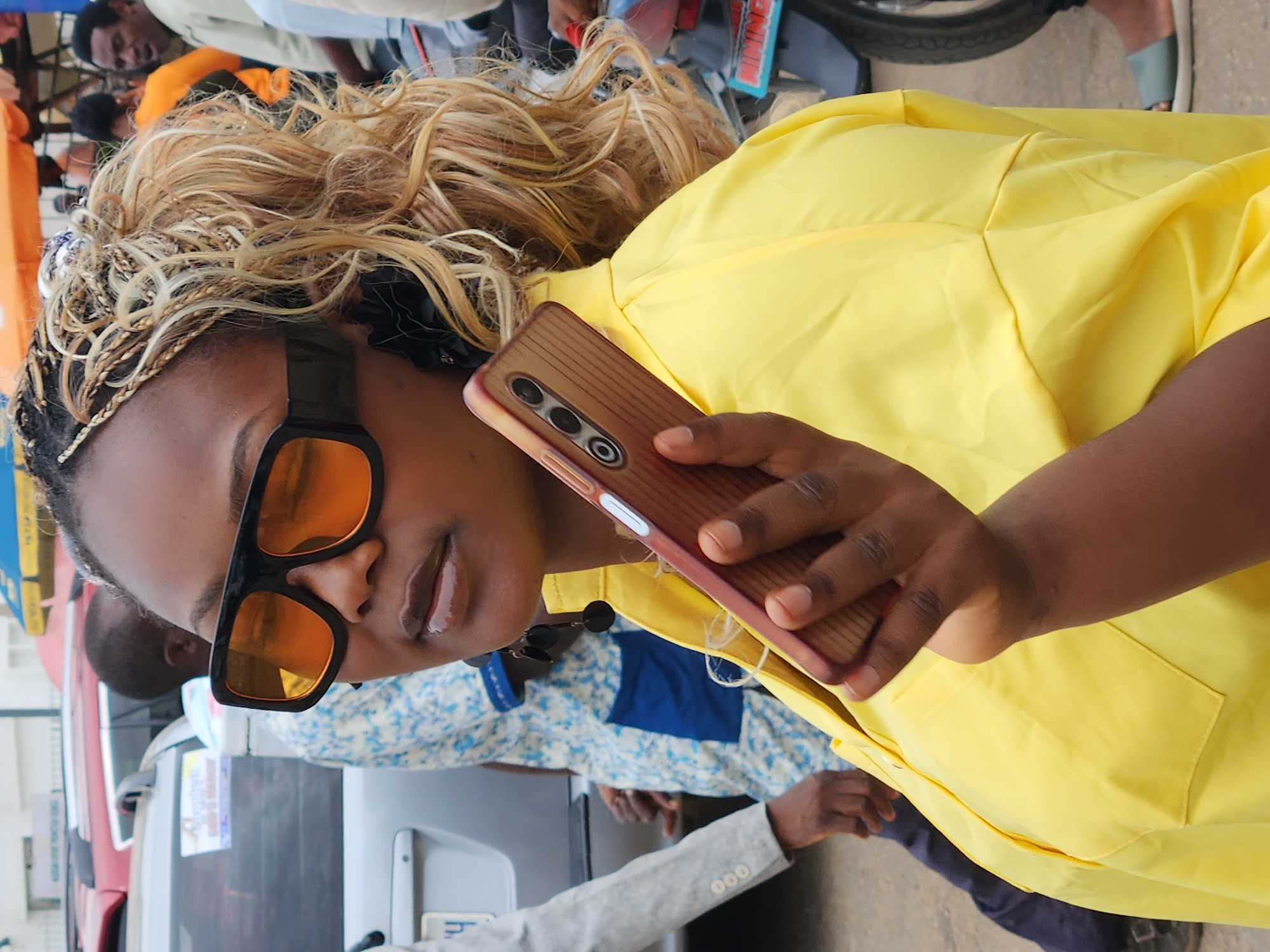 The media file shows a close-up of a woman in Jos, Nigeria, holding a smartphone. She is wearing a bright yellow shirt and large, black-framed sunglasses with orange lenses. Her hair is styled with blonde highlights and braided sections. In the background, other people are visible, and there are hints of vehicles, suggesting an outdoor setting, possibly a street or market. One person in the background is wearing an orange shirt. The woman appears to be looking down at her phone, with a neutral expression. The lighting suggests it is daytime. A portion of a gray blazer is visible on another individual in the foreground, and a blue and white patterned shirt is visible on someone behind the woman. There is no visible text in the frame.