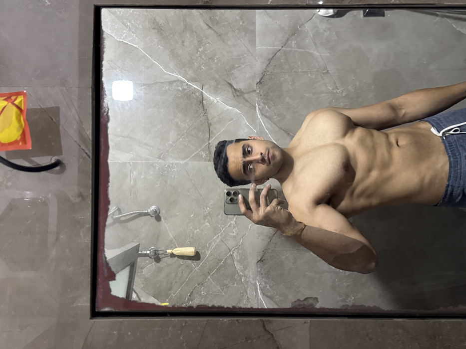 A muscular man stands shirtless in front of a bathroom mirror, taking a selfie with his phone. He's wearing dark blue shorts, and his torso is the focal point. The background includes gray marble tiles, a shower head, and a black-framed mirror with a red edge. A small abstract painting hangs on the wall to the left.