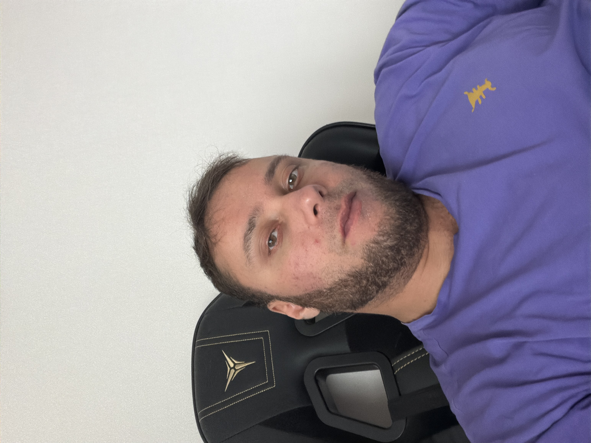 A man is seated in a black gaming chair wearing a purple long-sleeved shirt with a yellow logo on the upper right chest. He has short brown hair, light eyes, and a beard. The background is a plain white wall. The location is unknown and cannot be identified from the image.