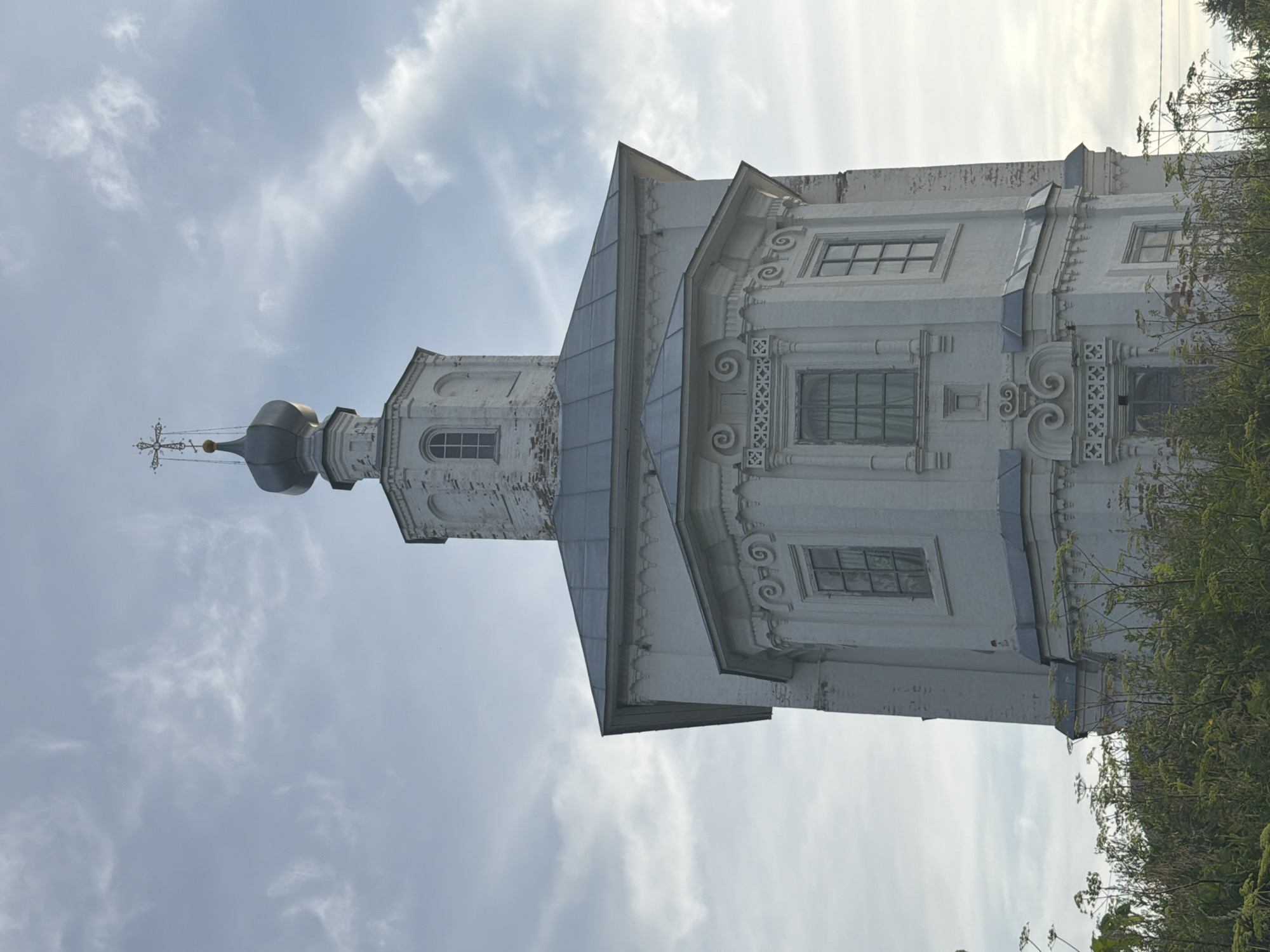 The image shows a close-up, low-angle view of a white, multi-tiered Orthodox church tower against a cloudy sky. The tower has octagonal sections with arched windows and ornate architectural details. A dark green dome with a cross tops the structure. In the foreground, dense green vegetation, including tall plants with umbrella-shaped flowers, obscures the lower part of the church. The scene is set in the city of Cherdyn, Russia.