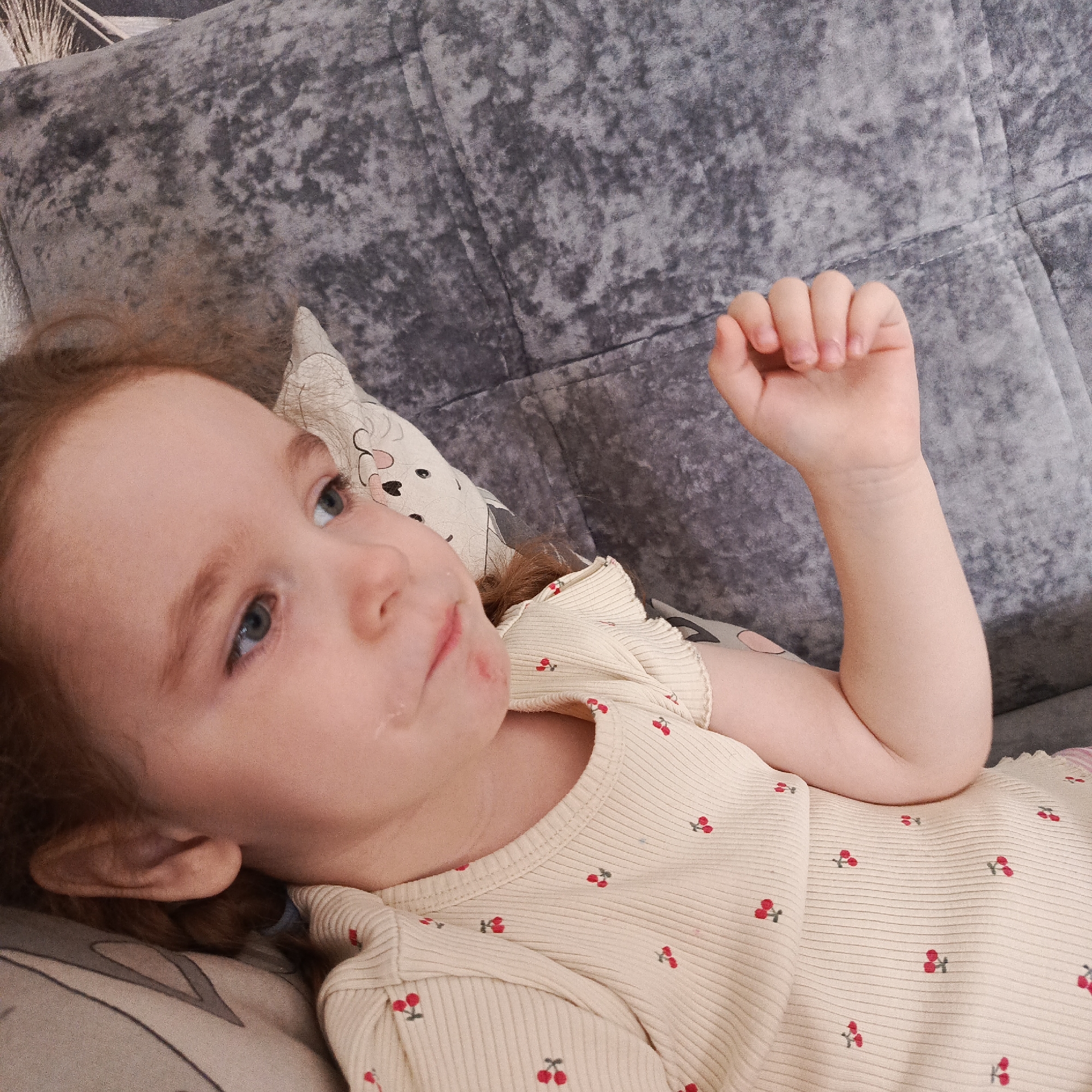 A toddler is lying on a gray couch or sofa. She is wearing a light yellow ribbed shirt with a small red cherry pattern. Her head rests on a pillow with a black and white design. She is looking upwards and her right hand is raised with her fingers slightly curled. The location is Марковский, Russia.